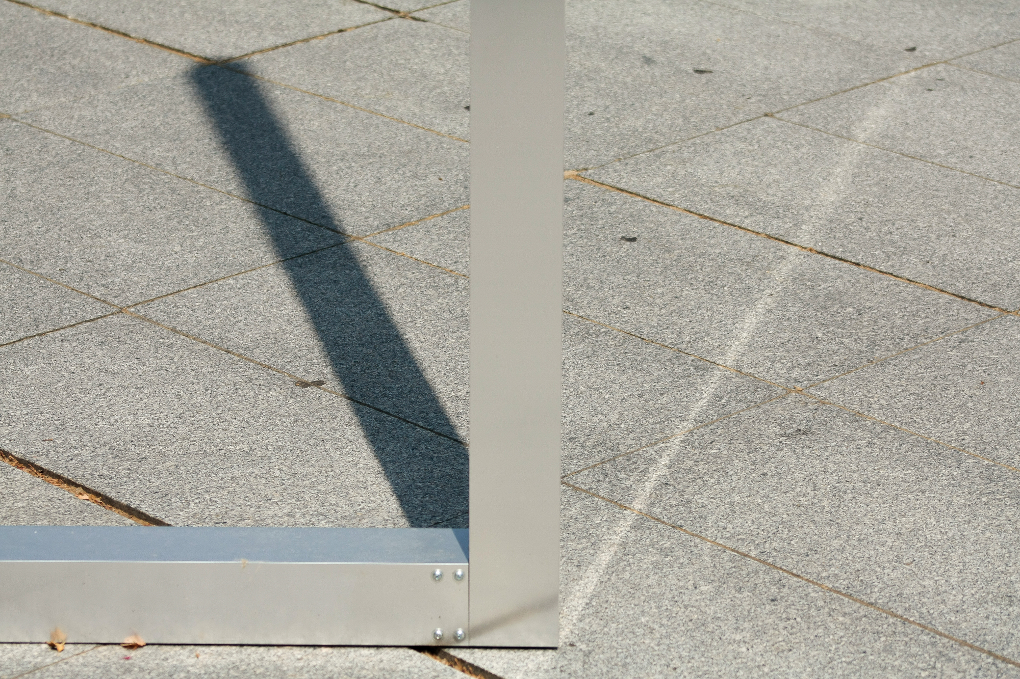 Close-up of a bolted aluminium corner joint on granite pavers, casting a long diagonal shadow in hard sunlight