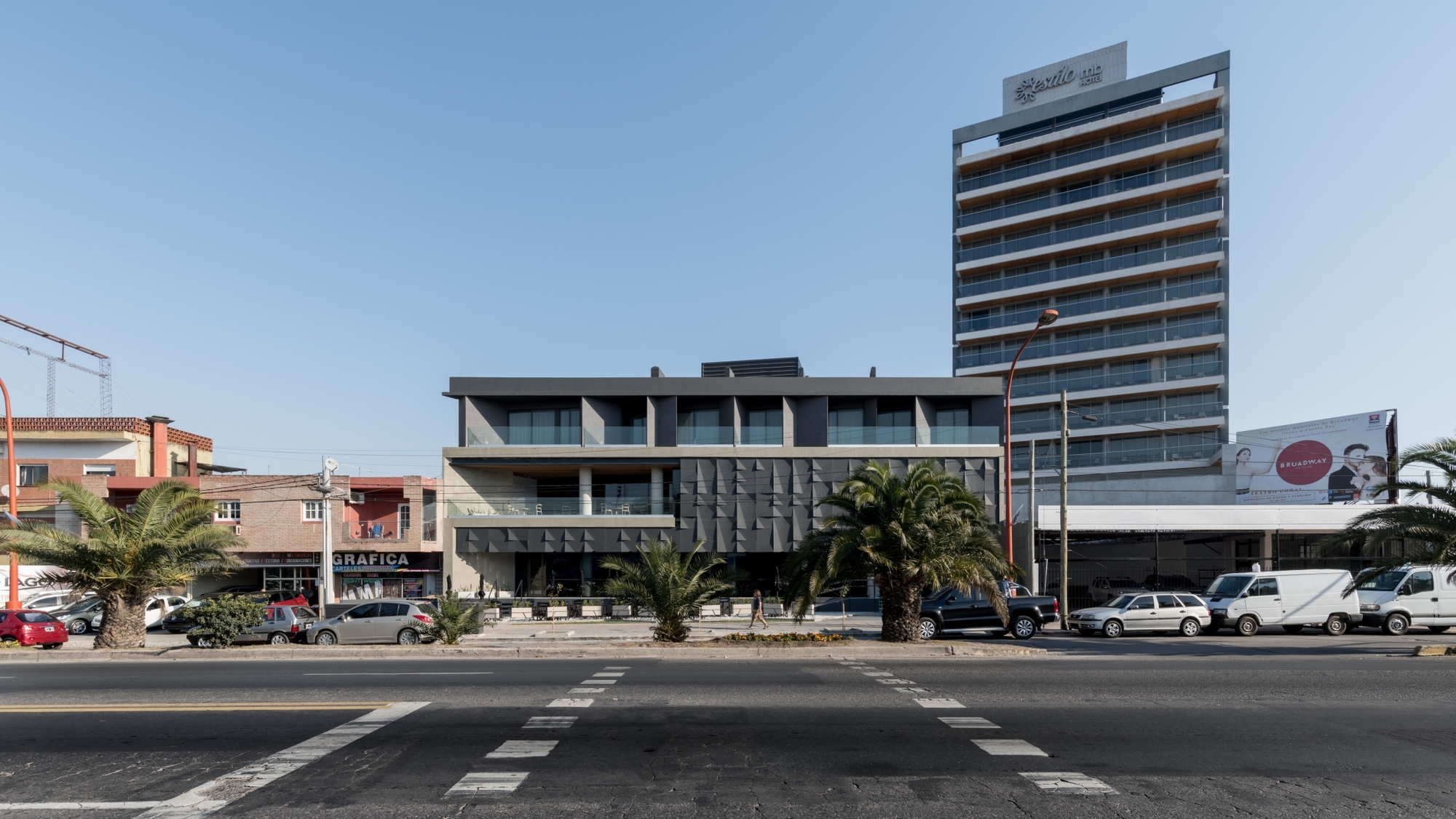 Street view of a low gray block beside a tall hotel tower, with palm trees and parked vans under clear blue sky