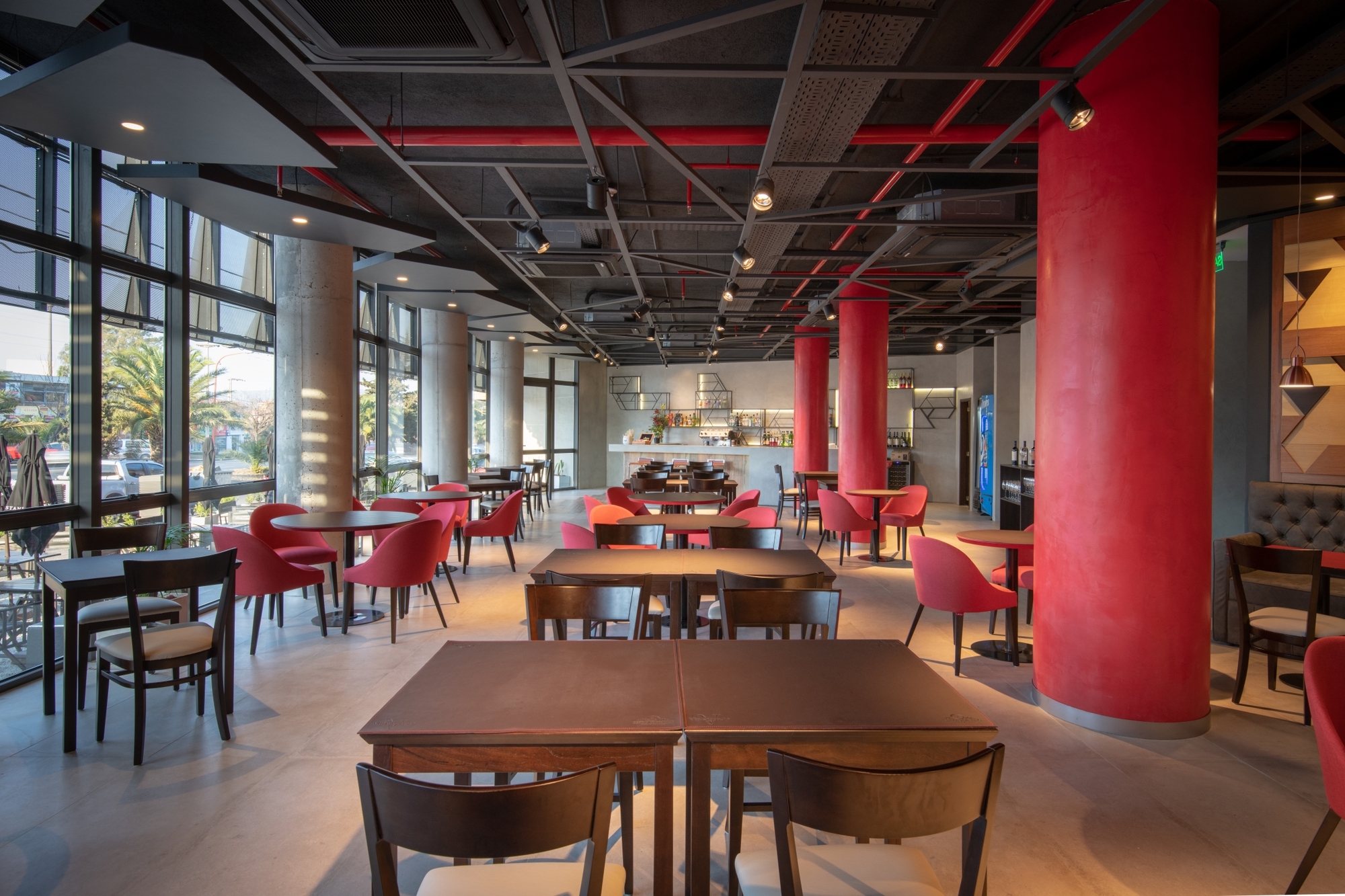 Restaurant interior with red round columns, dark wood tables, pink chairs and exposed ceiling ducts beside a glazed wall