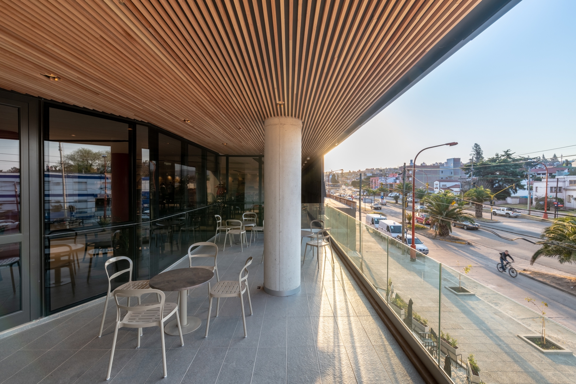 Covered terrace with a timber slat ceiling, concrete column and white chairs overlooking a street at low evening sun