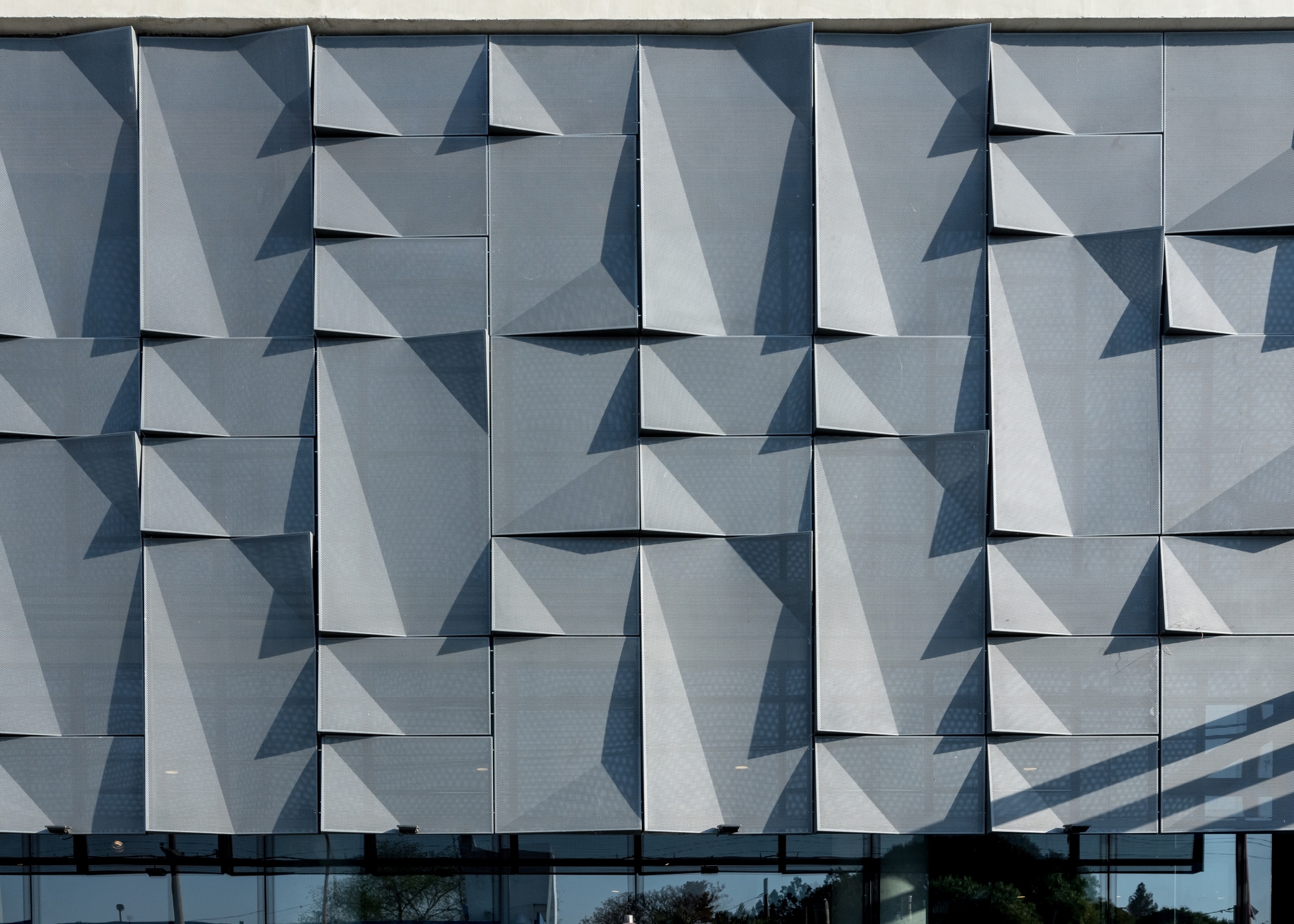 Close-up of the folded perforated metal facade panels casting angular shadows above a glazed ground floor