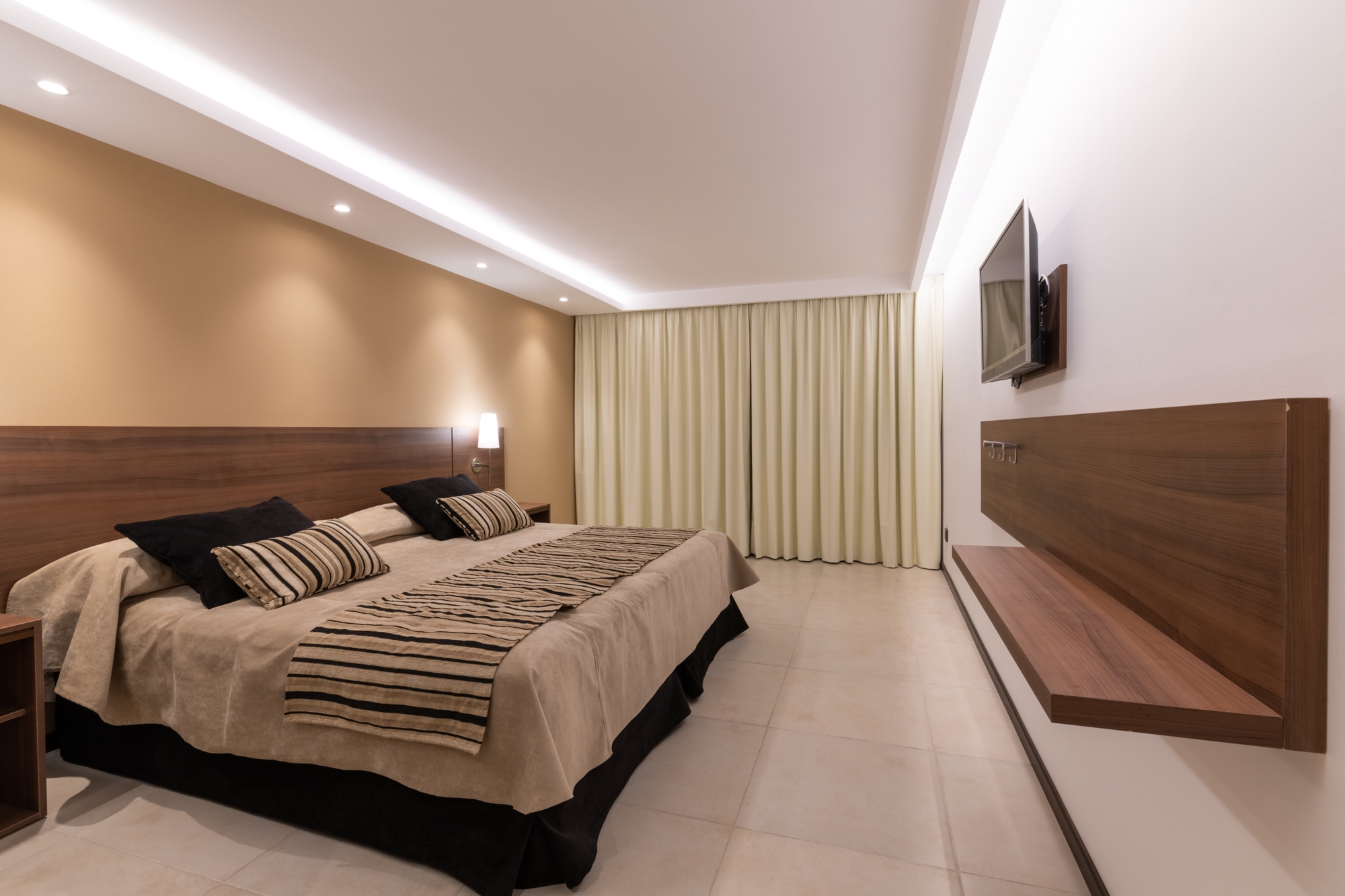 Hotel bedroom with a wood headboard, striped bed runner, floating wall shelf and cream curtains under a lit ceiling cove