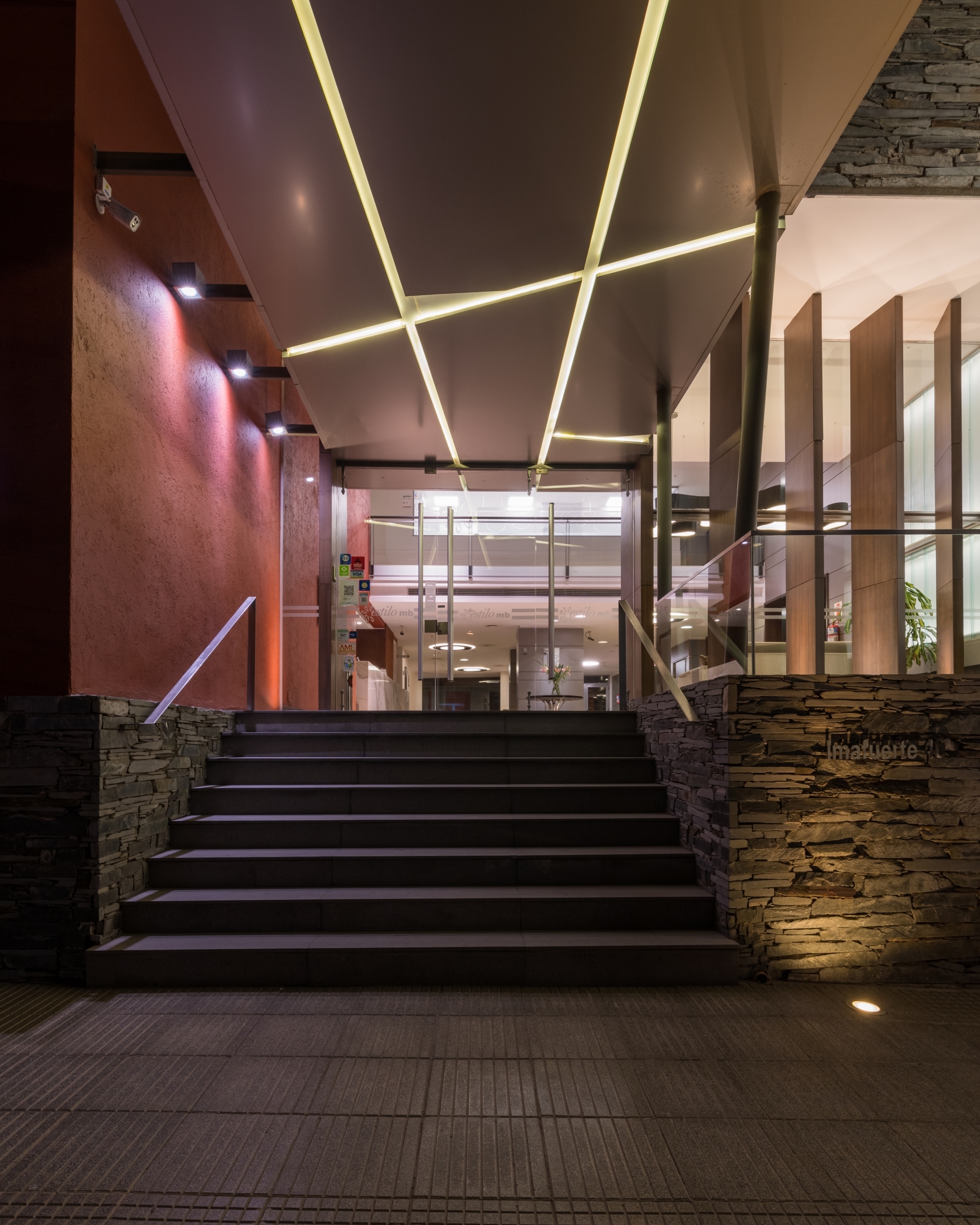 Night view of the entrance stair between slate-clad walls, lit by diagonal strip lights in the canopy ceiling