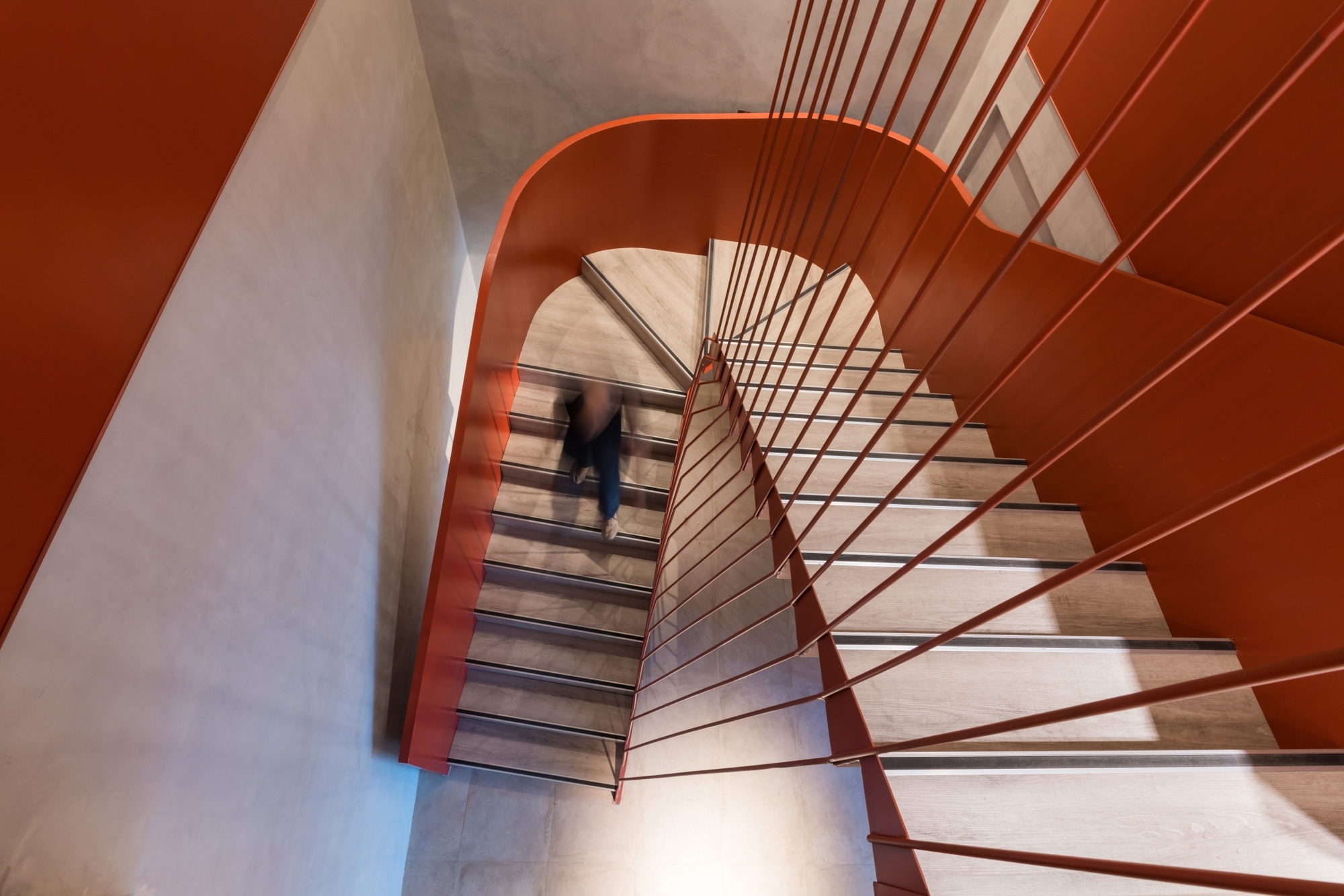 Top-down view of a rust-red steel staircase with thin vertical balusters and a blurred figure descending the treads