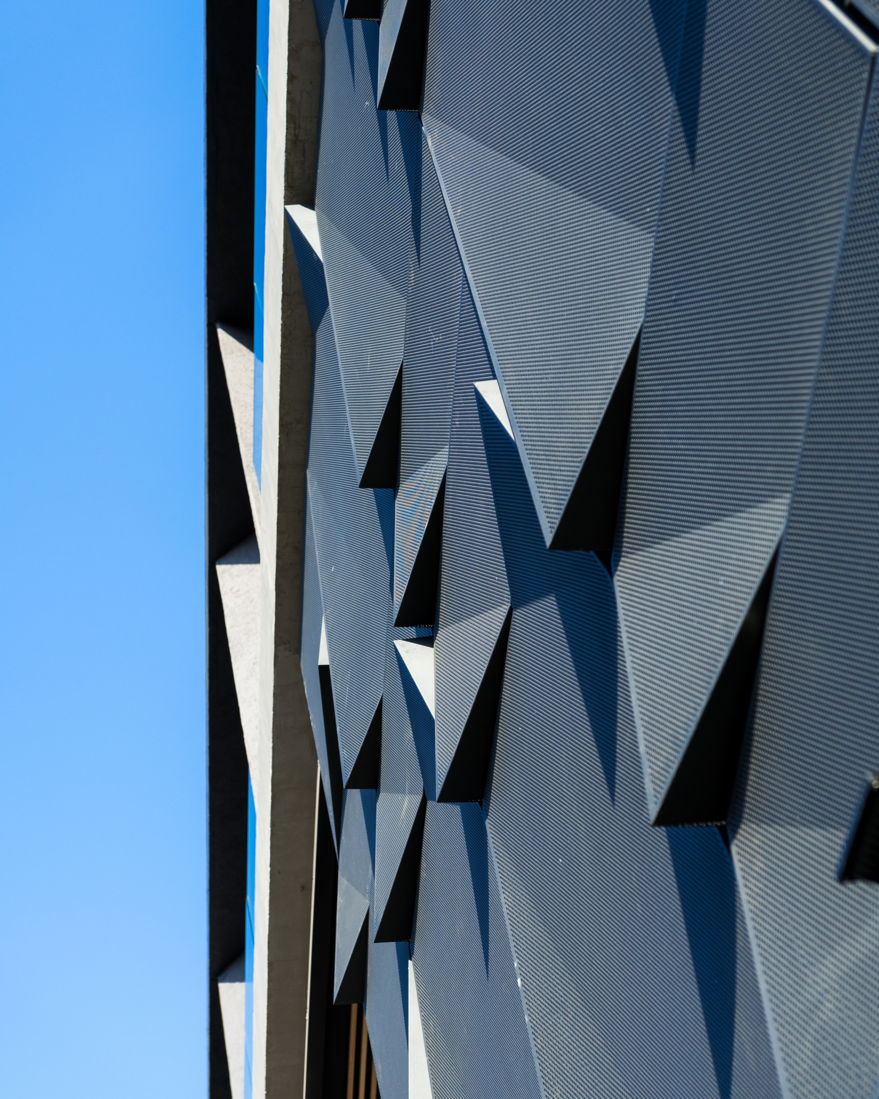 Close-up of faceted perforated metal facade panels beside a concrete edge under a clear blue sky