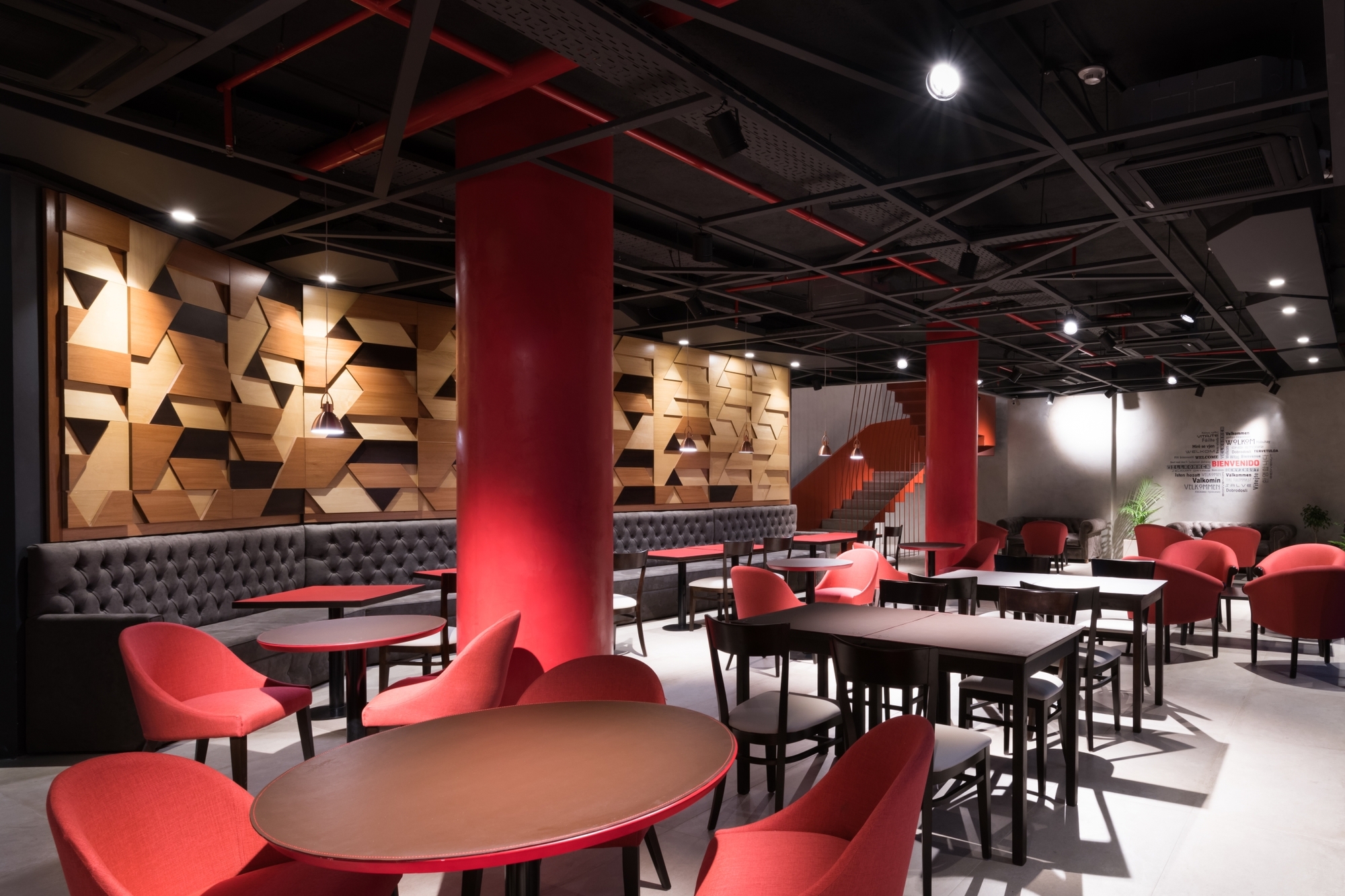 Restaurant interior with red columns, red armchairs, tufted banquette and a faceted timber wall panel under a dark exposed ceiling