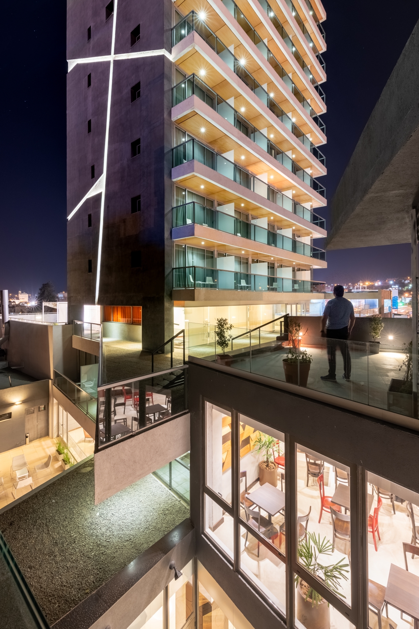 Night view of a residential tower with glass balconies and lit soffits, a man standing on a terrace above a cafe