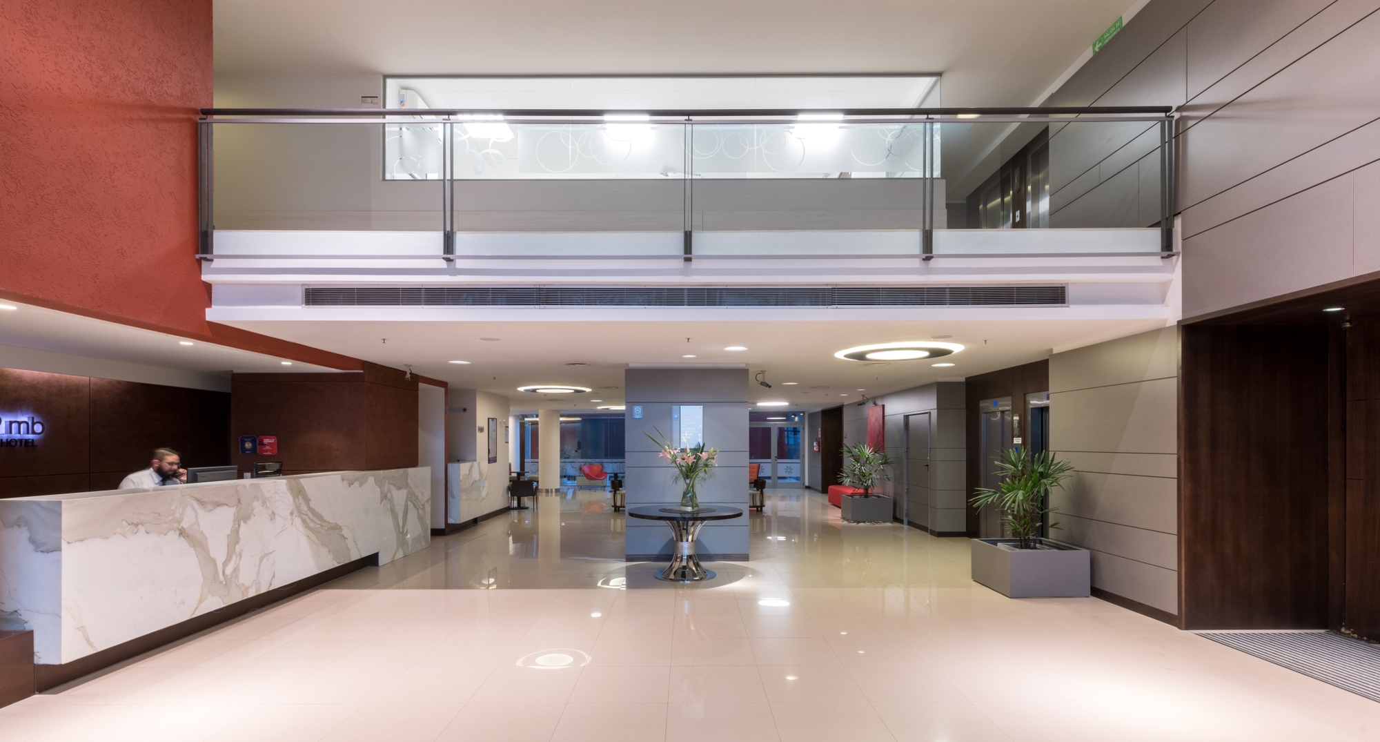Double-height lobby with marble reception counter, glass-balustrade mezzanine, potted plants and a flower table on a polished floor