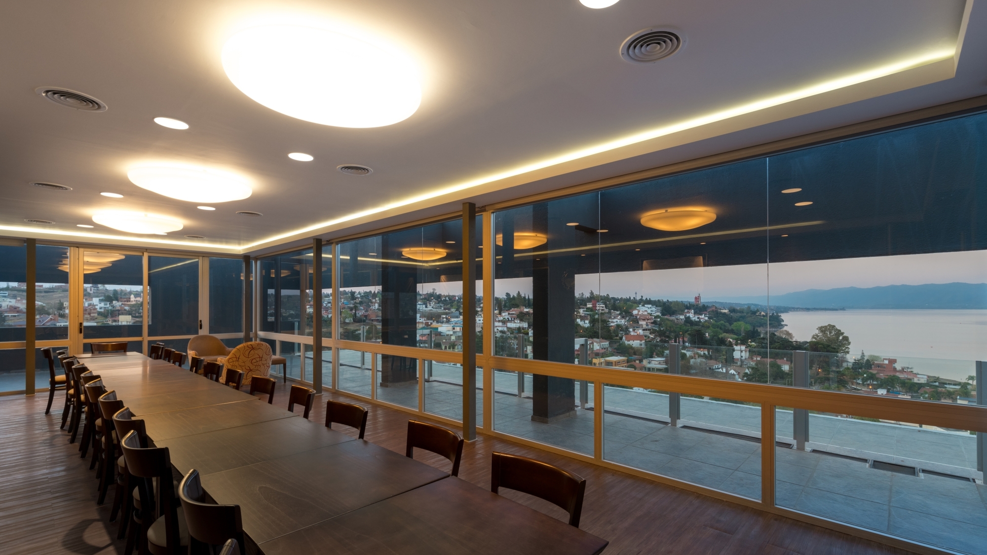 Long dining table and wooden chairs beside a glazed wall with timber mullions, looking out over a lake town at dusk