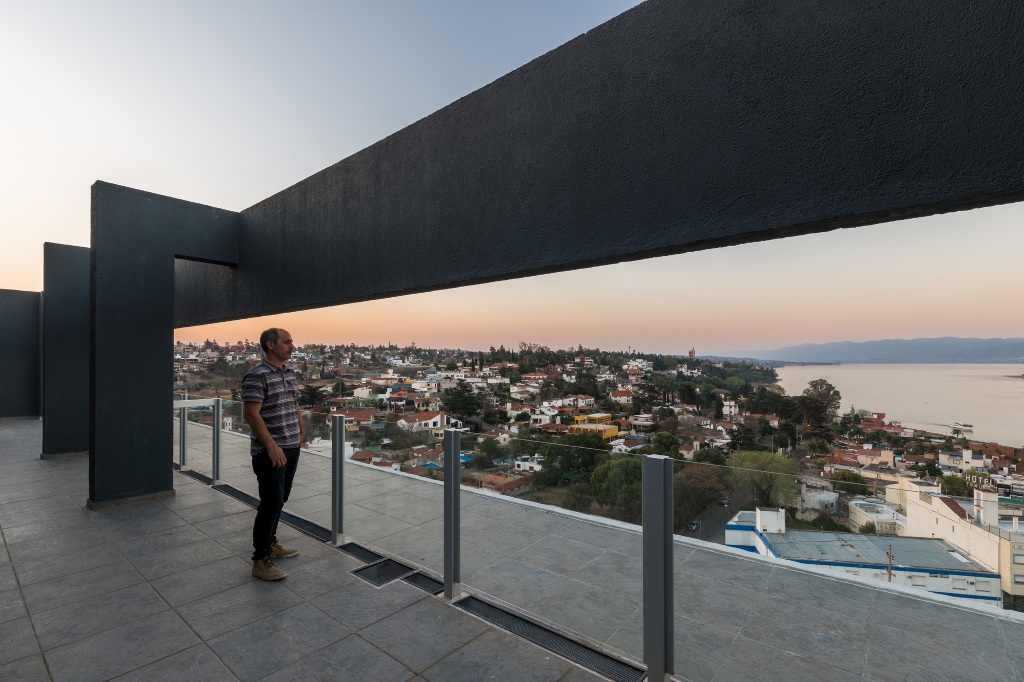 Man standing on a tiled terrace beneath a dark concrete frame, looking over rooftops and a lake at sunset