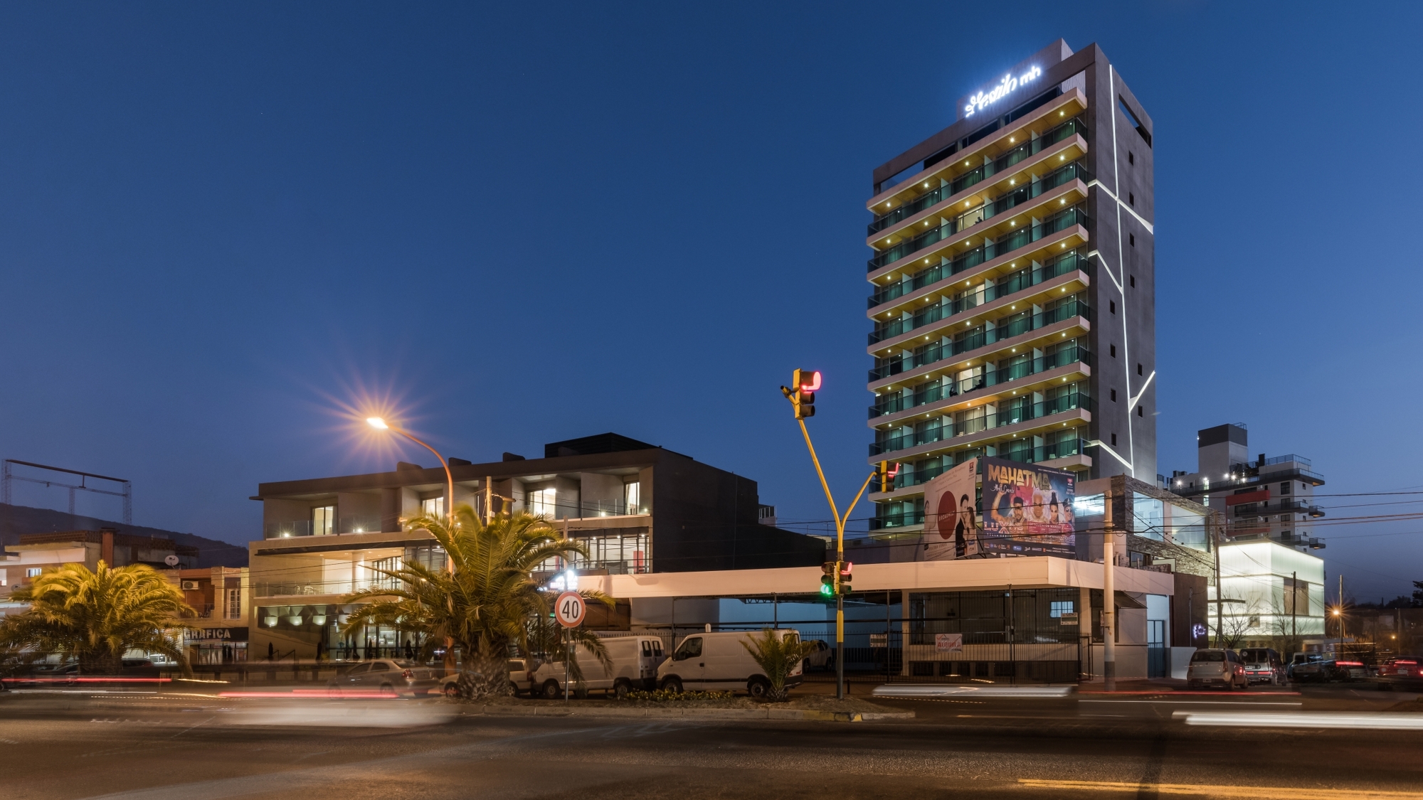 Tall hotel tower with stacked balconies and a lit white line on its facade, seen from a street with light trails at dusk