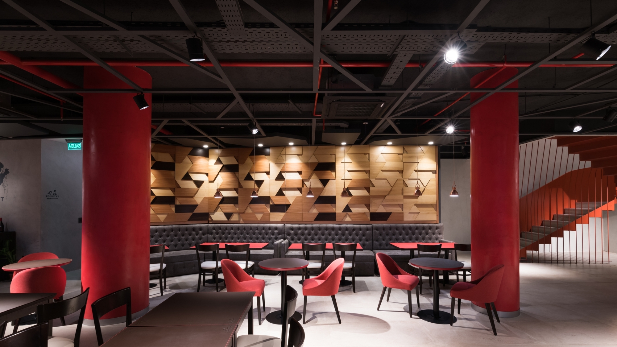Restaurant interior with two red round columns, a faceted timber wall behind a grey banquette and a red staircase, under a black ceiling
