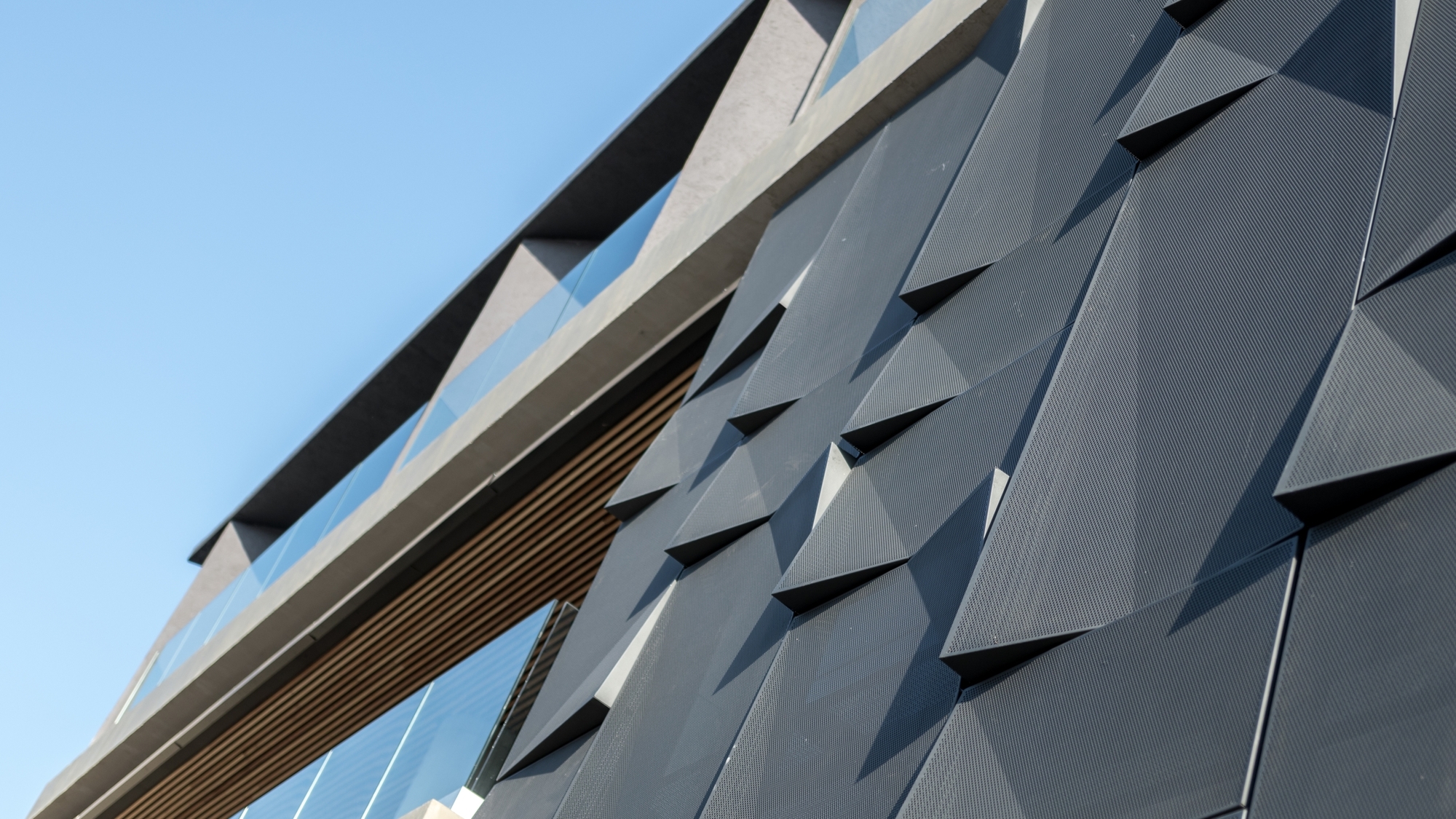Upward view of the faceted dark metal cladding panels beside a timber soffit and glass balustrade under a clear blue sky