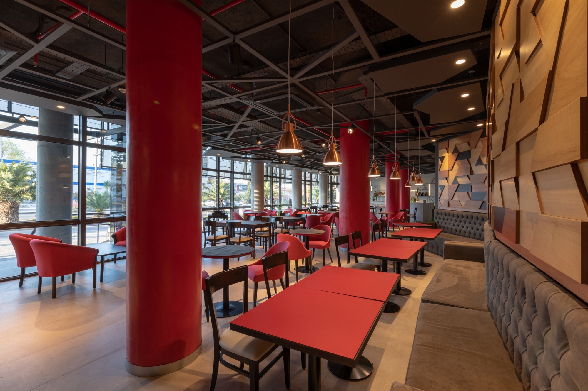 Café dining room with red tables and chairs, red columns and copper pendant lamps, with palm trees visible through the glazing