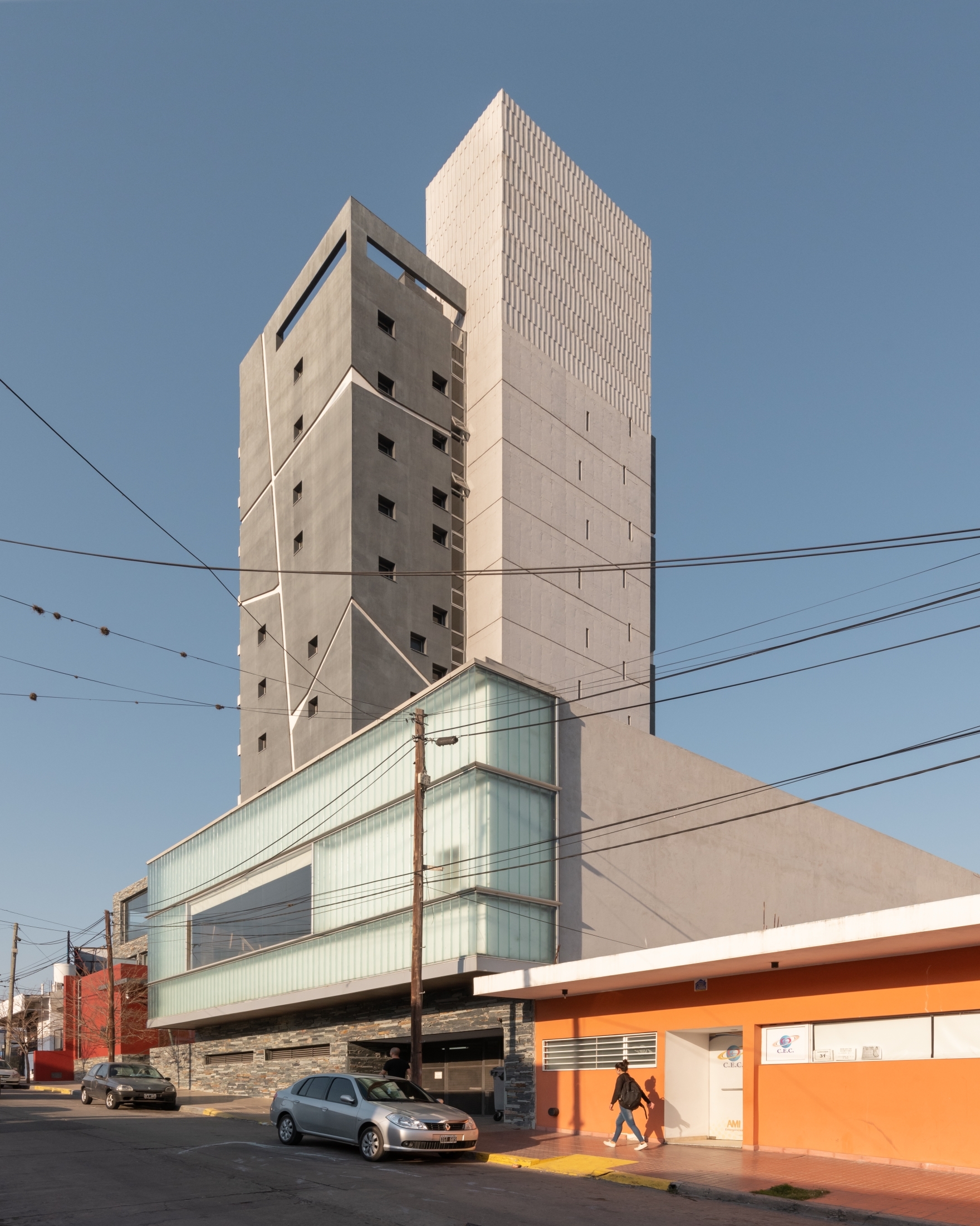 Street corner view of a faceted concrete tower above a glass-clad base, with a pedestrian passing an orange building under clear sky