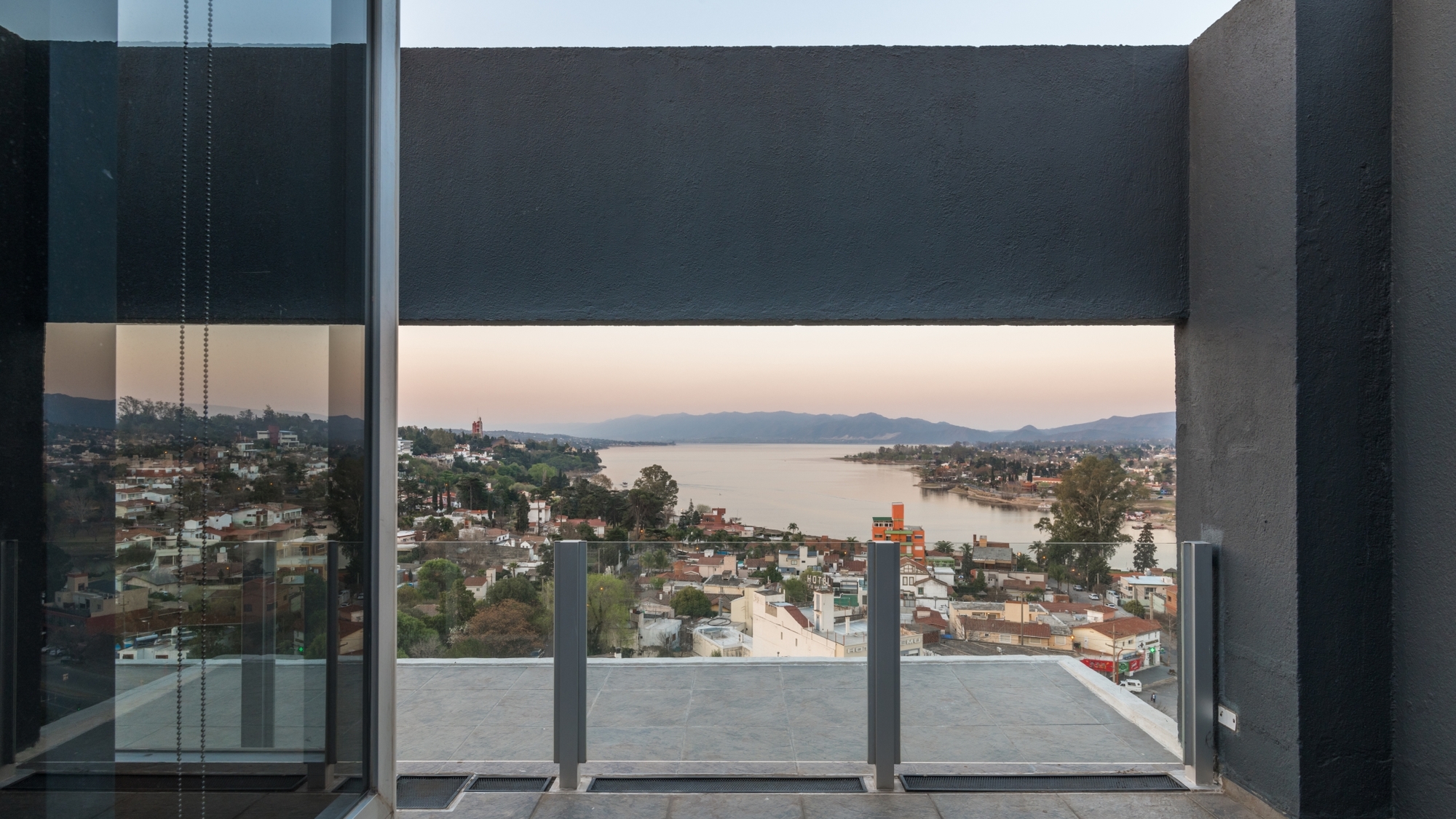 View through a dark concrete frame and glass balustrade toward a lake, hills and town roofs at dusk