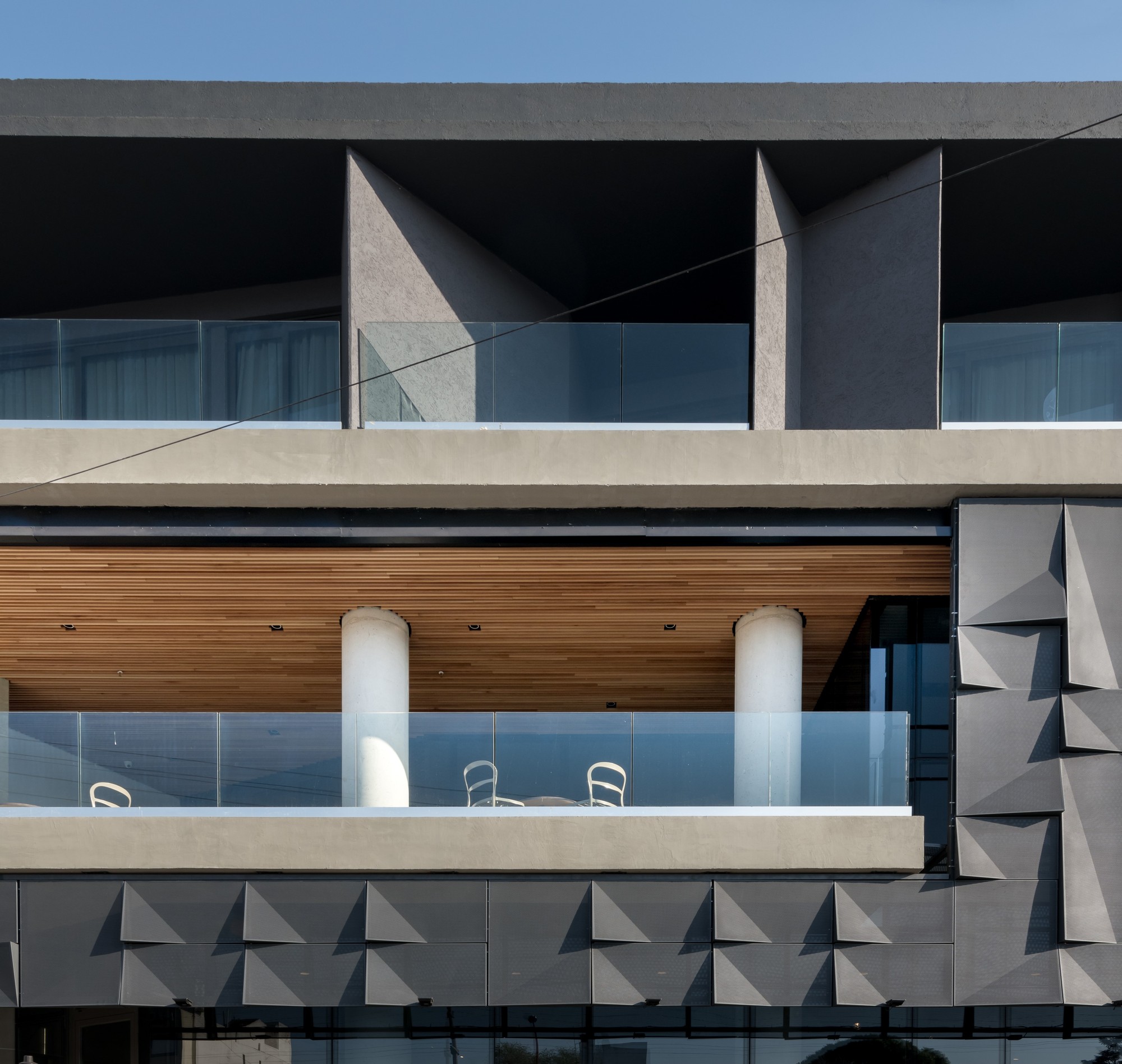 Facade detail of terraces with glass balustrades, round white columns, a timber soffit and faceted grey panels under blue sky