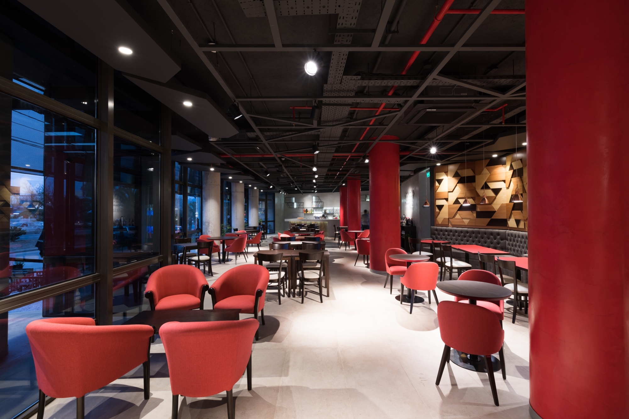 Restaurant interior with red columns, coral armchairs, exposed black ceiling and a faceted timber wall panel at night