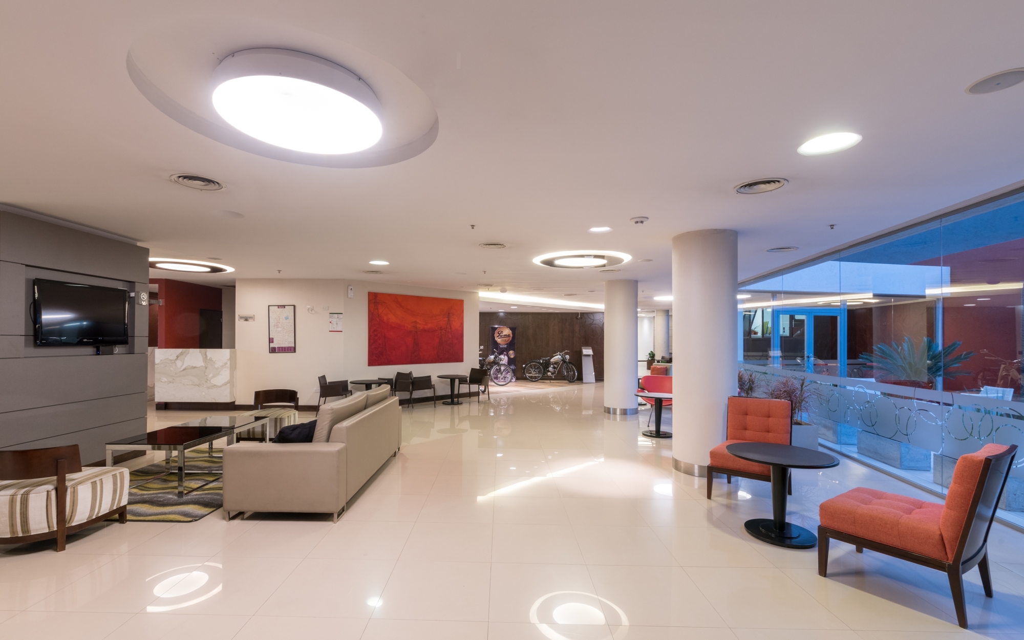 Hotel lobby with polished white floor, round ceiling lights, beige sofa and orange armchairs beside a glazed wall