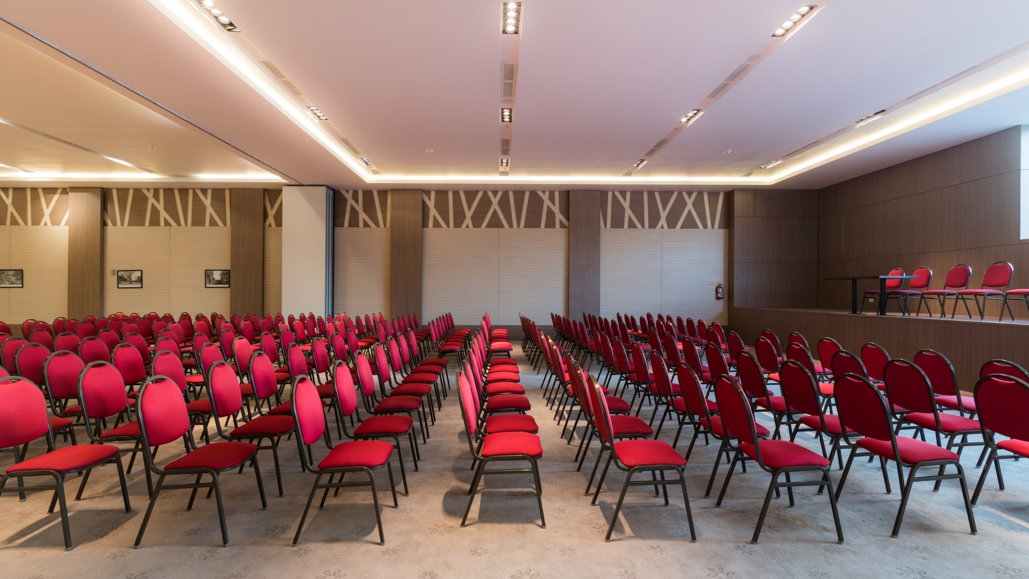 Conference room with rows of red upholstered chairs facing a low stage, lit by a continuous glowing ceiling cove