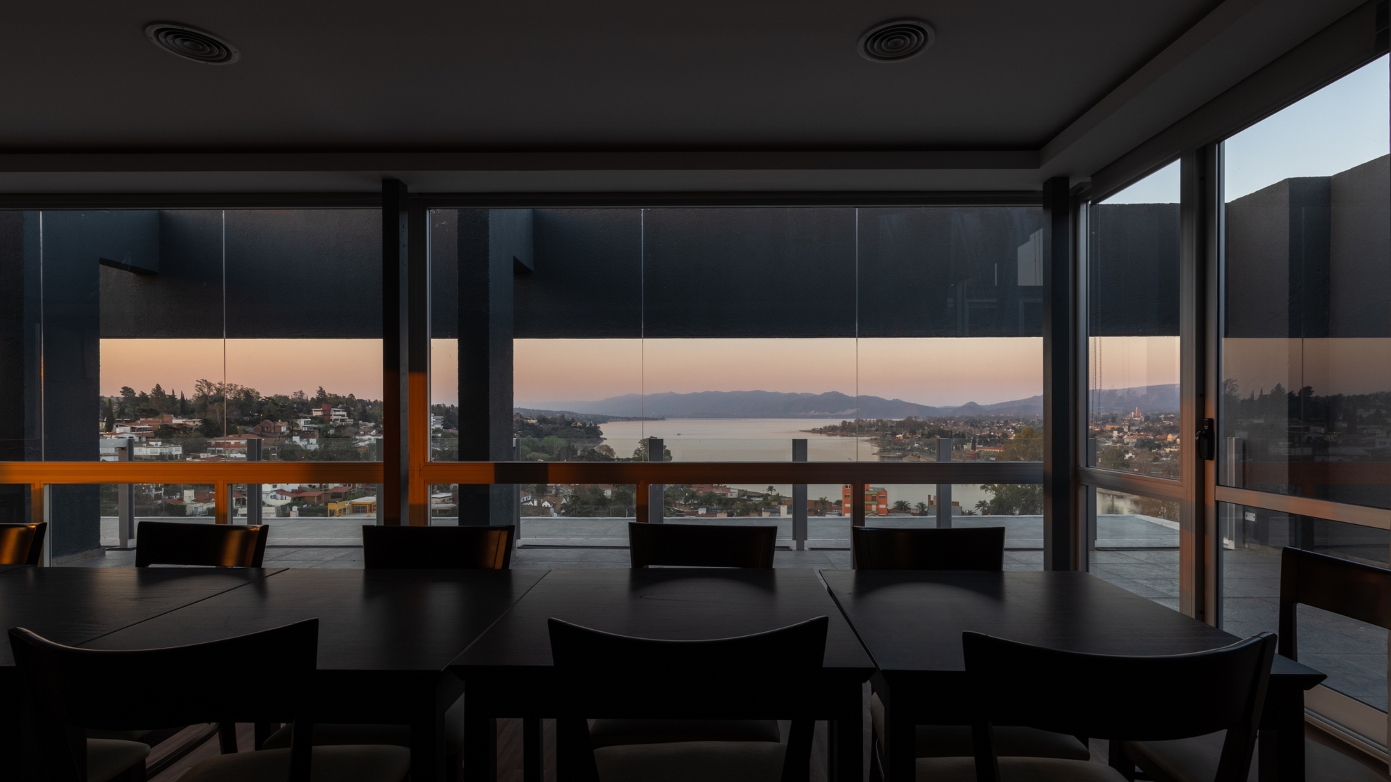 Dining tables and dark wood chairs facing floor-to-ceiling glazing with a lake and hills at sunset