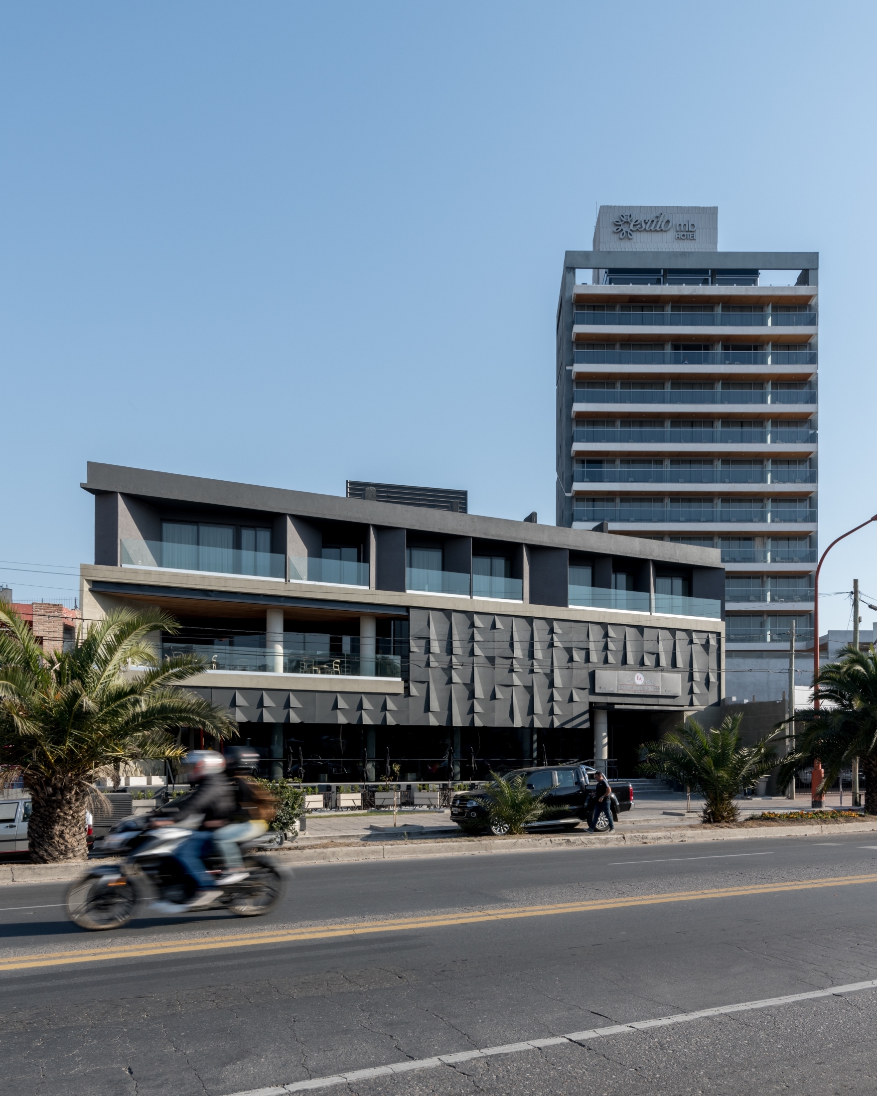 Street view of the low faceted dark panel facade with a tall tower behind and a motorcycle passing in blur