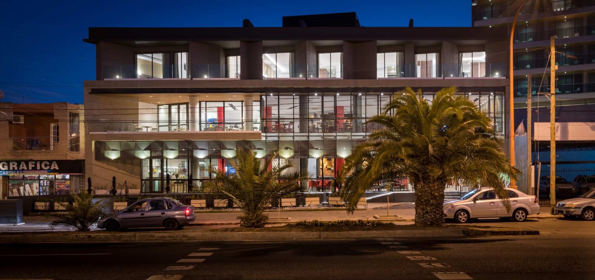 Night view of the lit glazed ground floor and terraces behind palm trees, with parked cars along the street