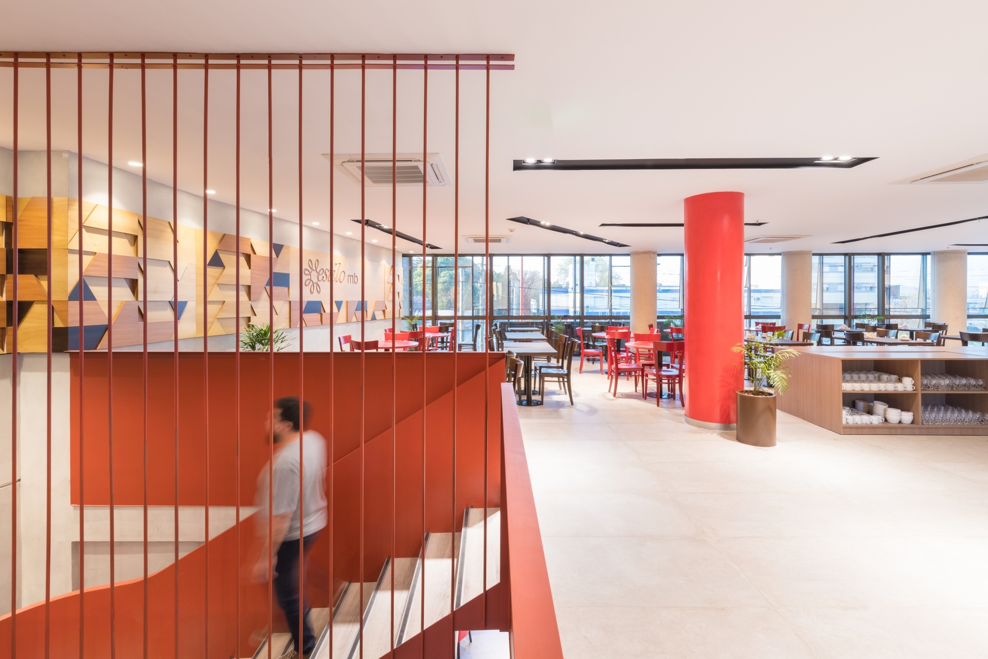 Rust-red stair with vertical rod screen overlooking a dining hall, a blurred figure descending beside a red column