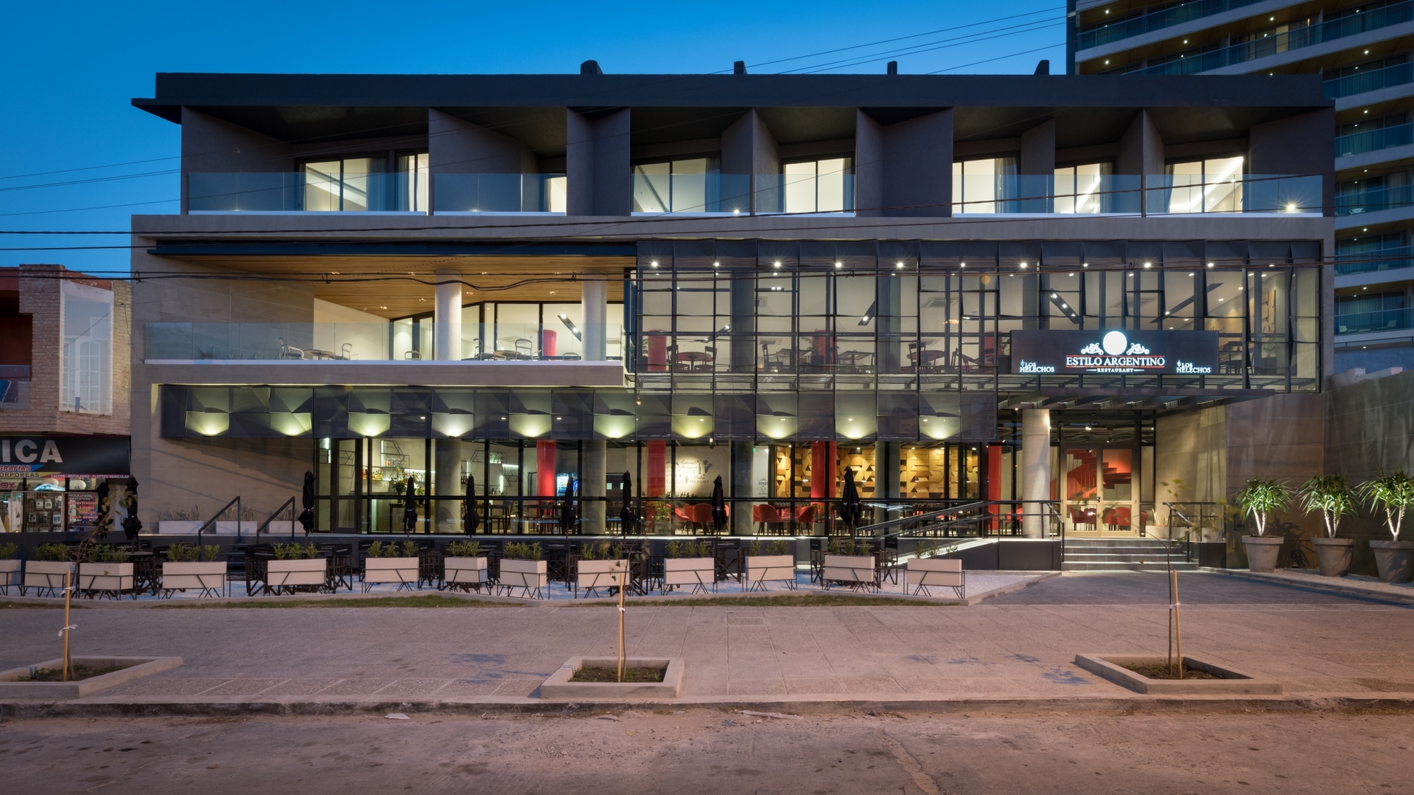Street-level facade of a glazed restaurant podium beneath dark upper floors, lit at blue hour with planters along the sidewalk