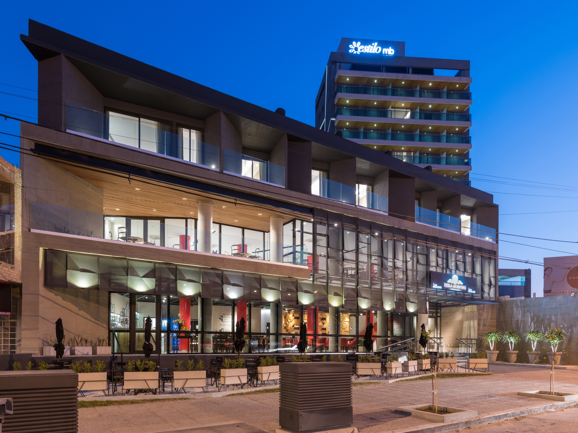 Corner view of the stepped glass and stone building with a hotel tower behind, glowing at dusk