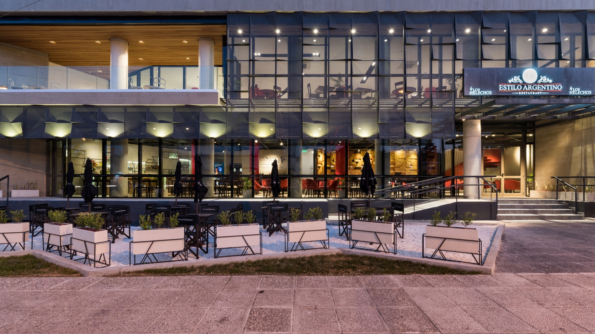 Ground-floor restaurant frontage with faceted glass soffit lights, planter boxes on white gravel and a lit entrance sign at dusk