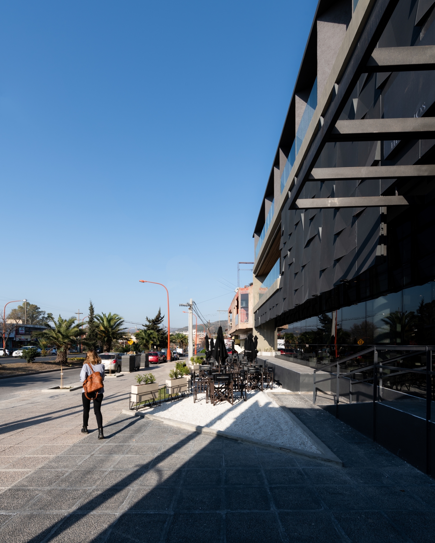 Pedestrian walking along the sidewalk beside the dark faceted facade, with café tables on white gravel under a clear blue sky