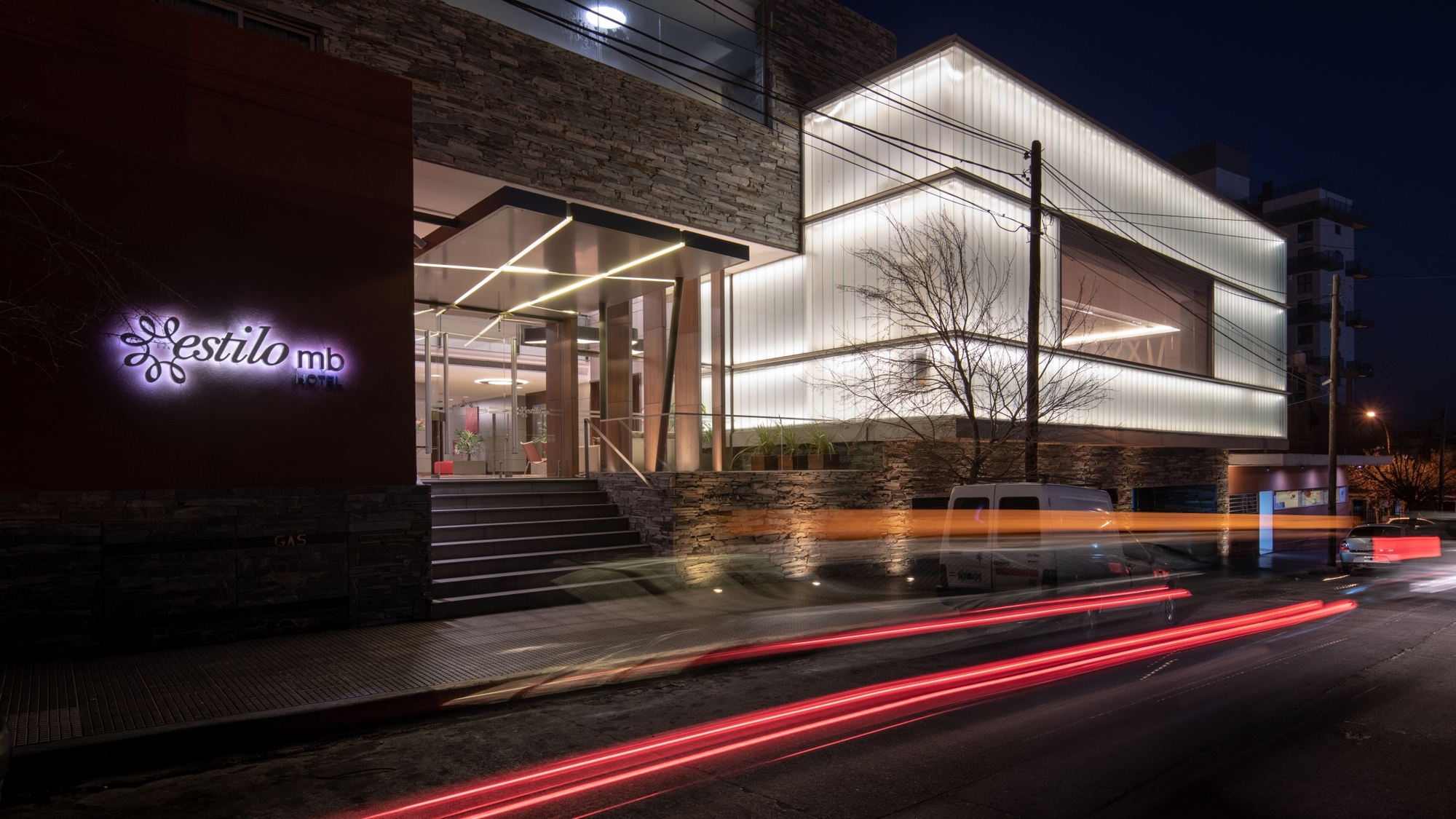 Translucent glowing glass box beside a stone-clad entrance stair, with red car light trails streaking across the street at night