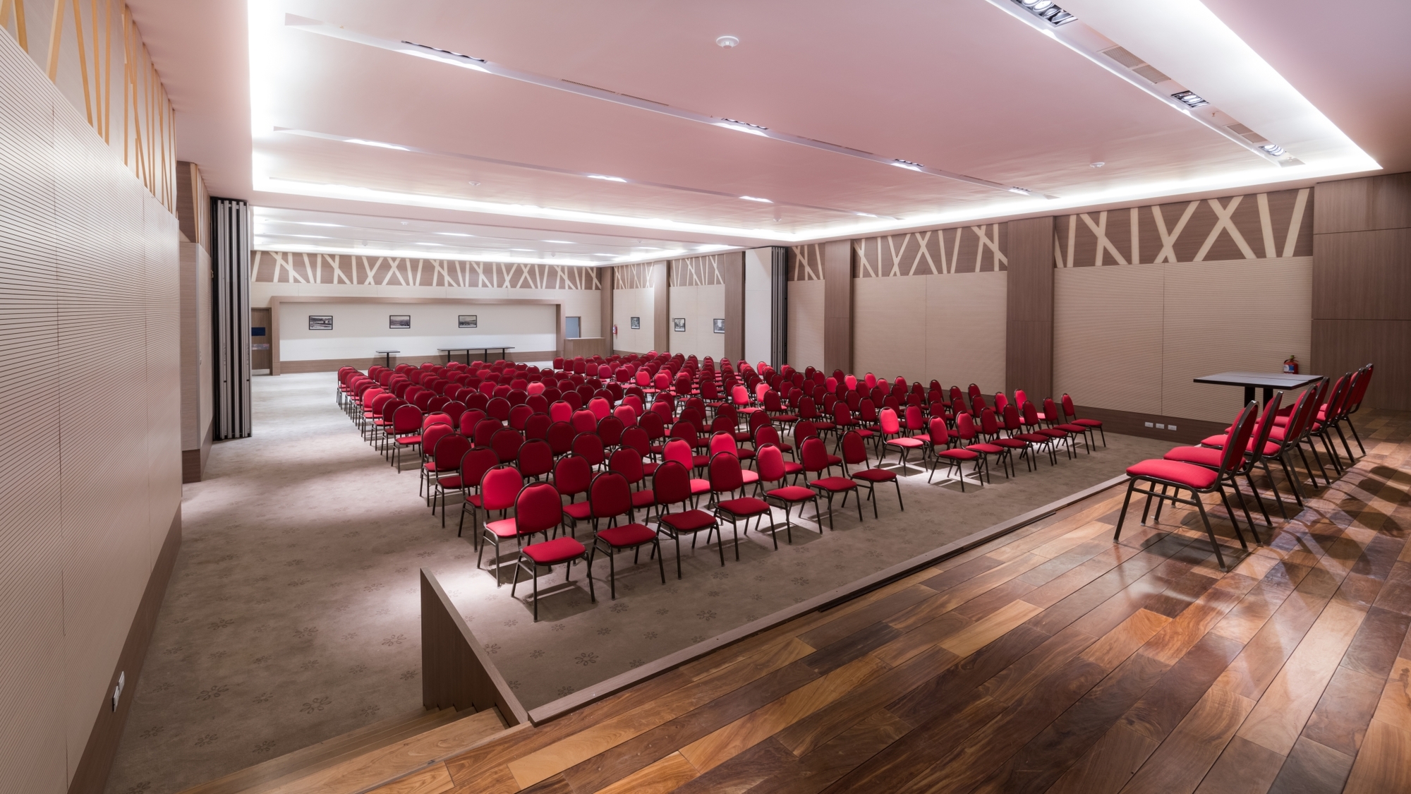 Conference hall with rows of red chairs on a carpeted platform, timber floor in the foreground and lit ceiling coves