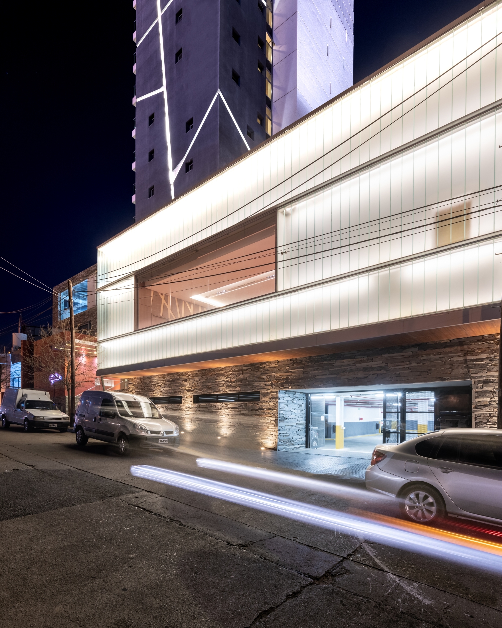 Night view of the translucent glowing facade above a stone-clad base and garage entrance, with a light trail from a passing car