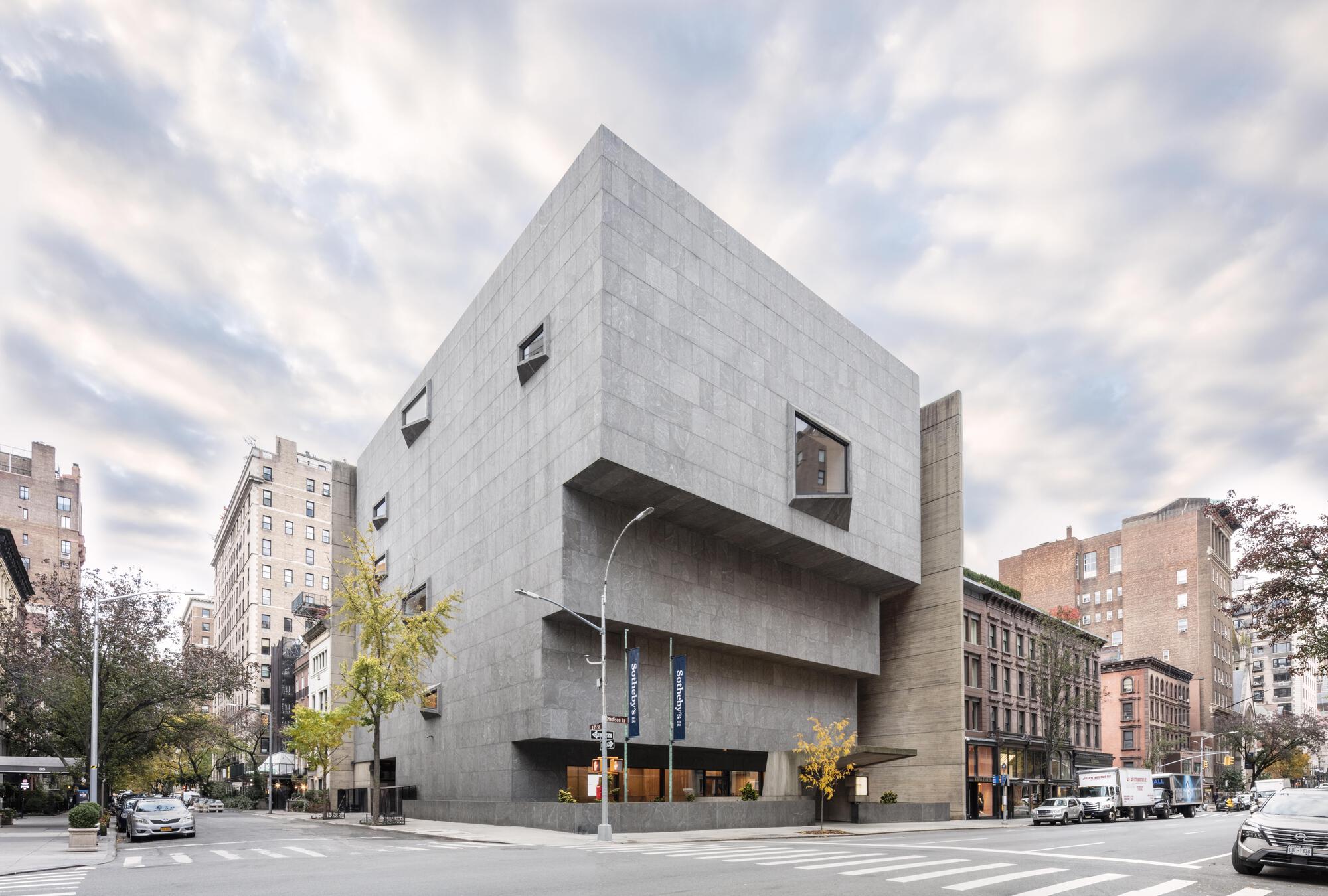 Corner view of the stepped granite-clad block with angled windows under a cloudy sky, street trees in autumn color