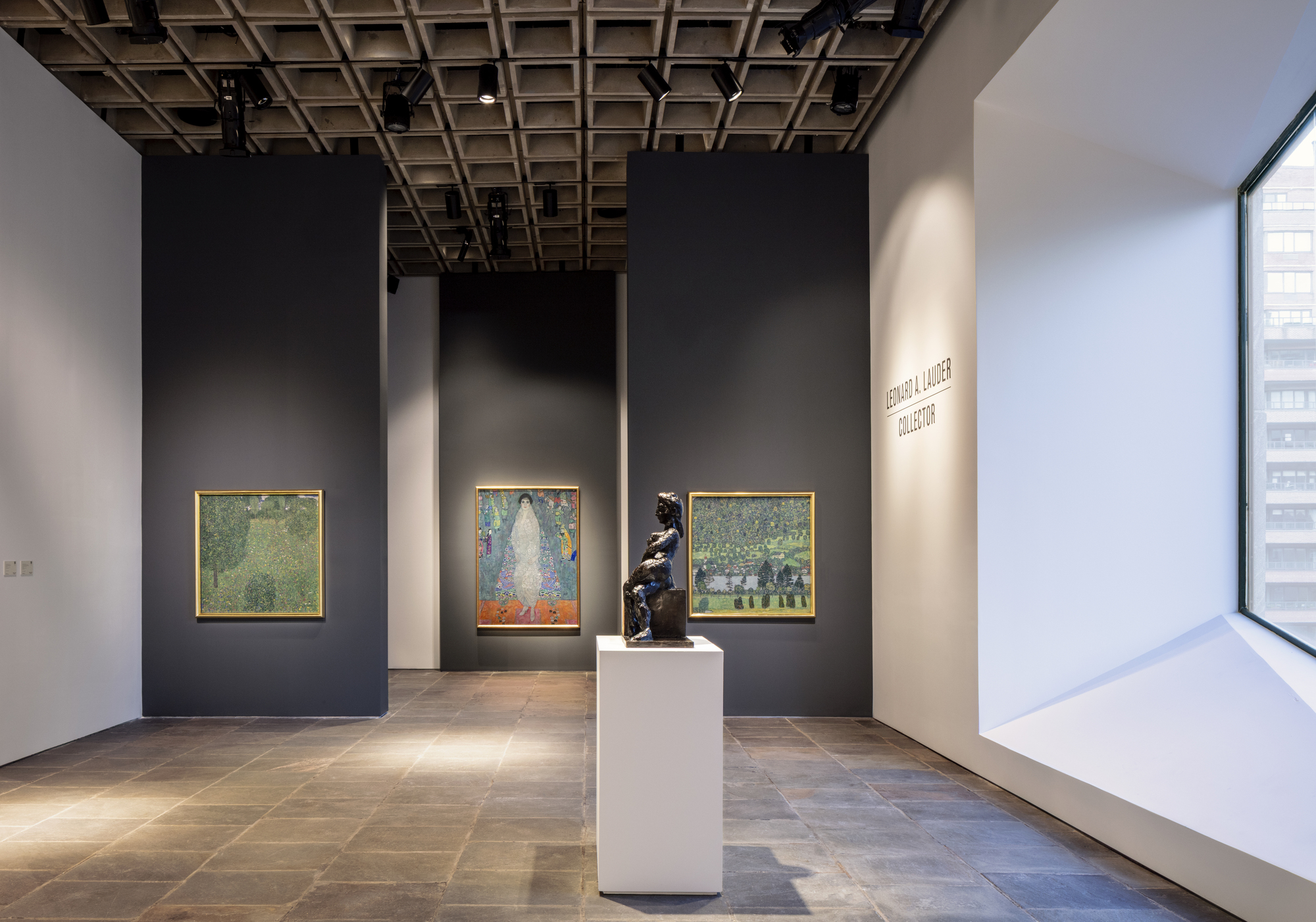 Gallery with charcoal partition walls, a bronze figure on a white plinth and a deep angled window frame letting in daylight