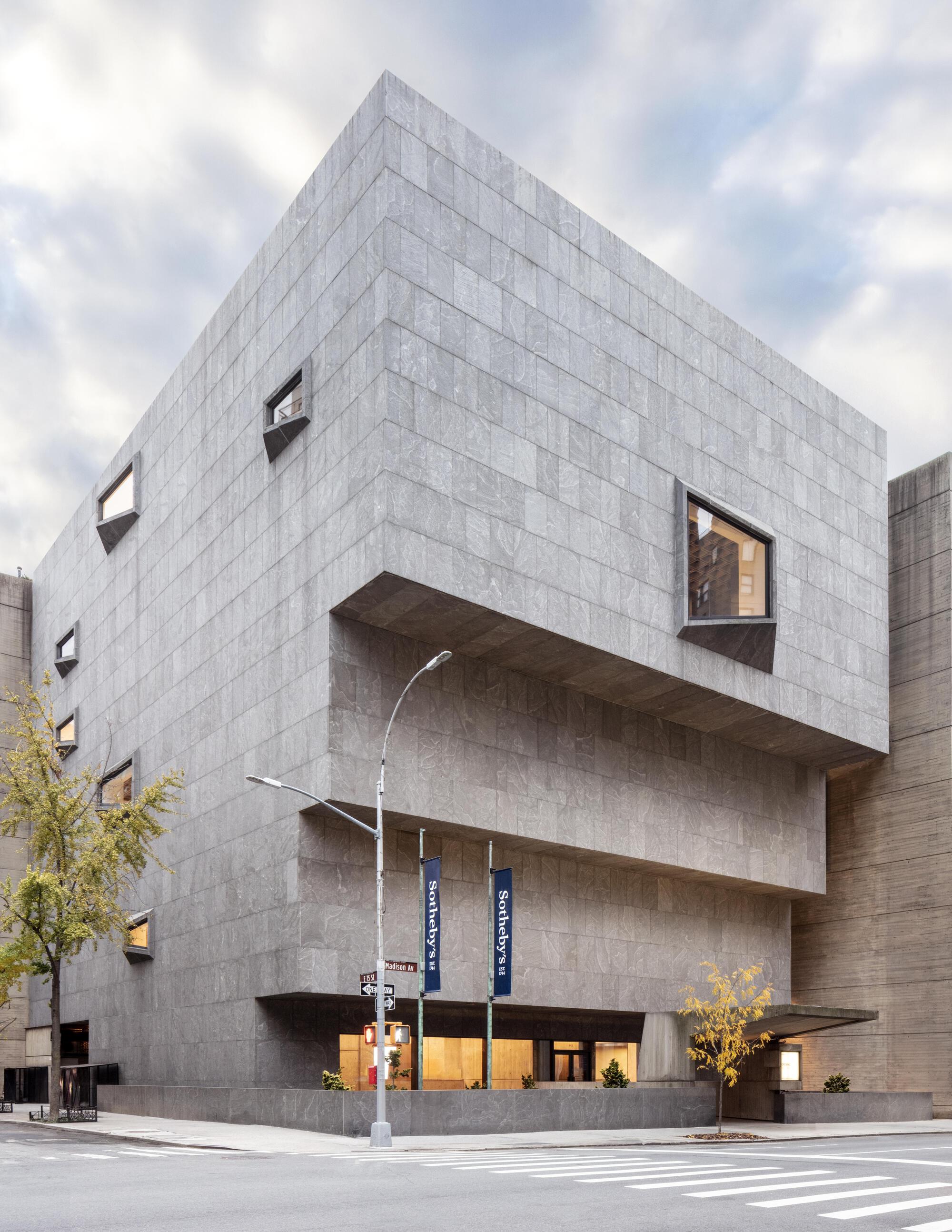 Vertical view of the gray stone facade with cantilevered upper volume, banners and a yellow-leaved tree at street level