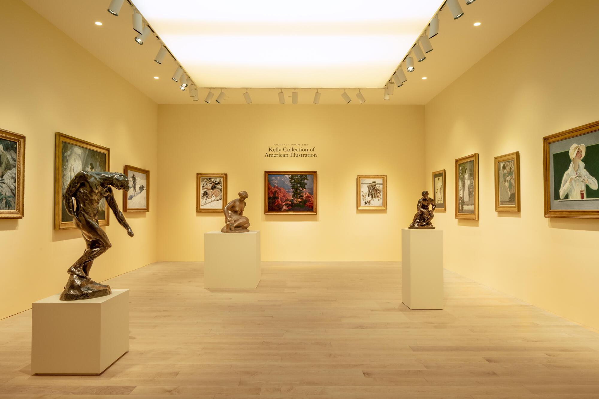 Gallery with pale yellow walls, bronze sculptures on plinths and gilt-framed illustrations under a glowing skylight panel and track lights