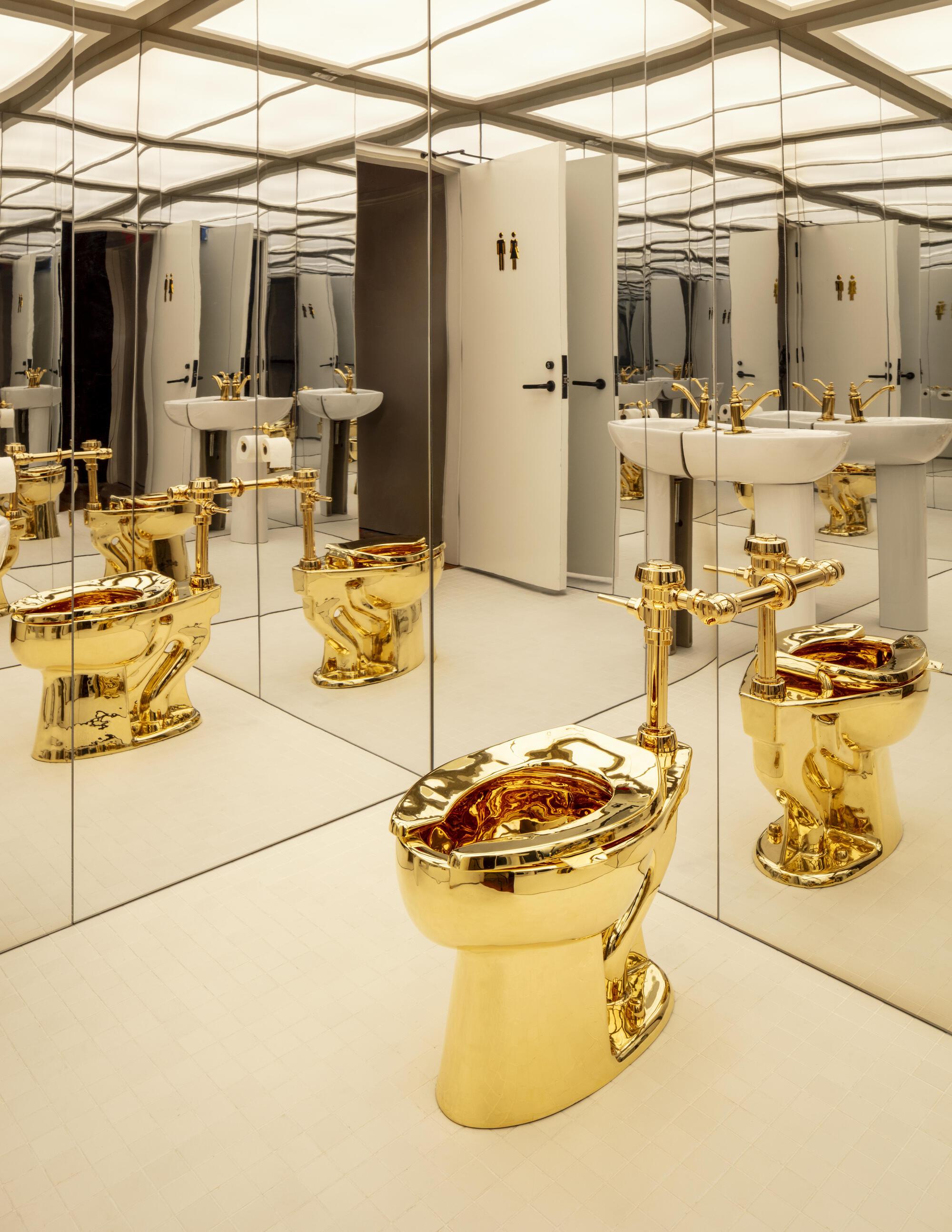 Gold toilet with brass piping in a mirrored restroom, repeated in reflections beside white pedestal sinks under a glowing ceiling