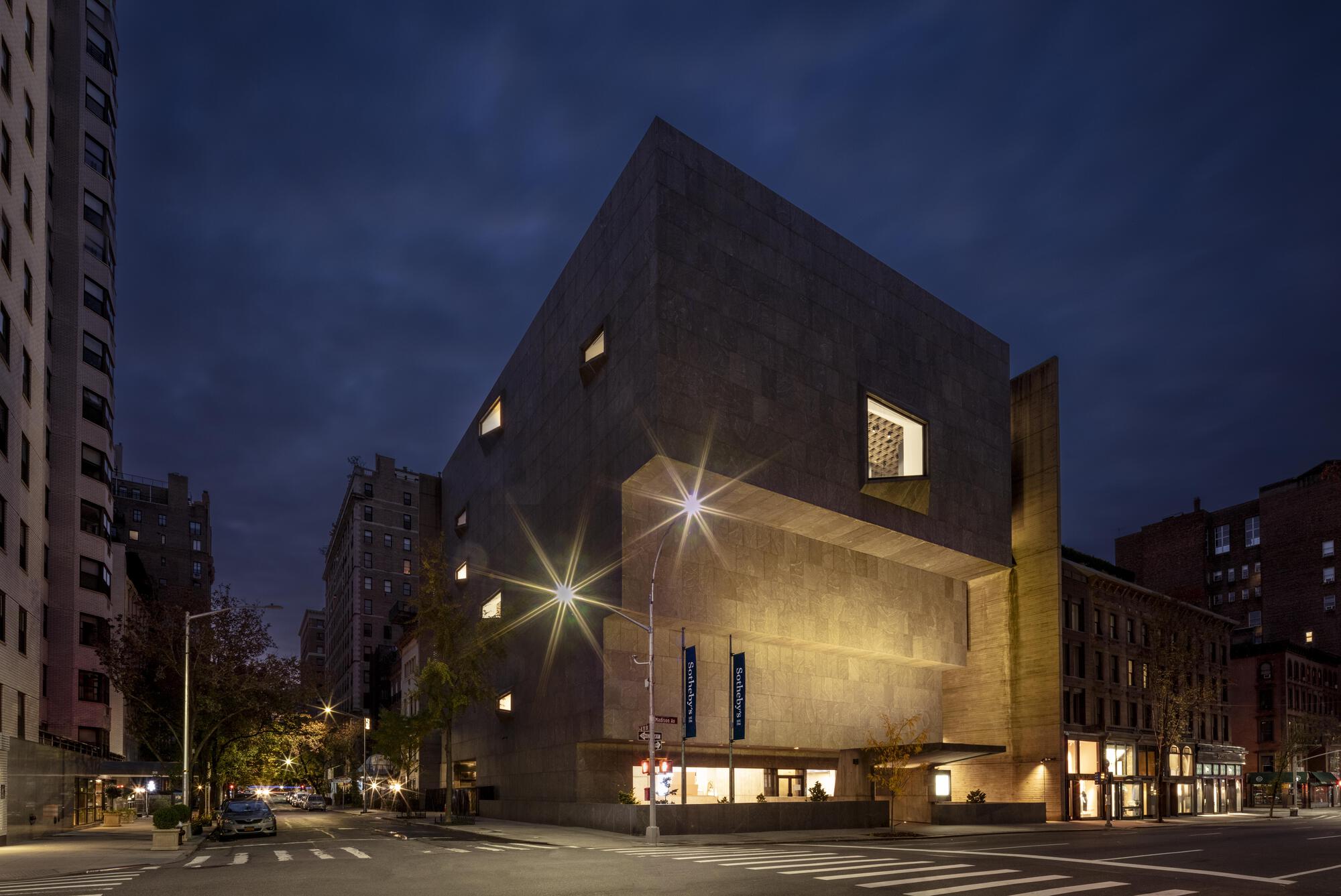 Corner view of the stepped granite building with angled window openings, lit by street lamps under a dark dusk sky