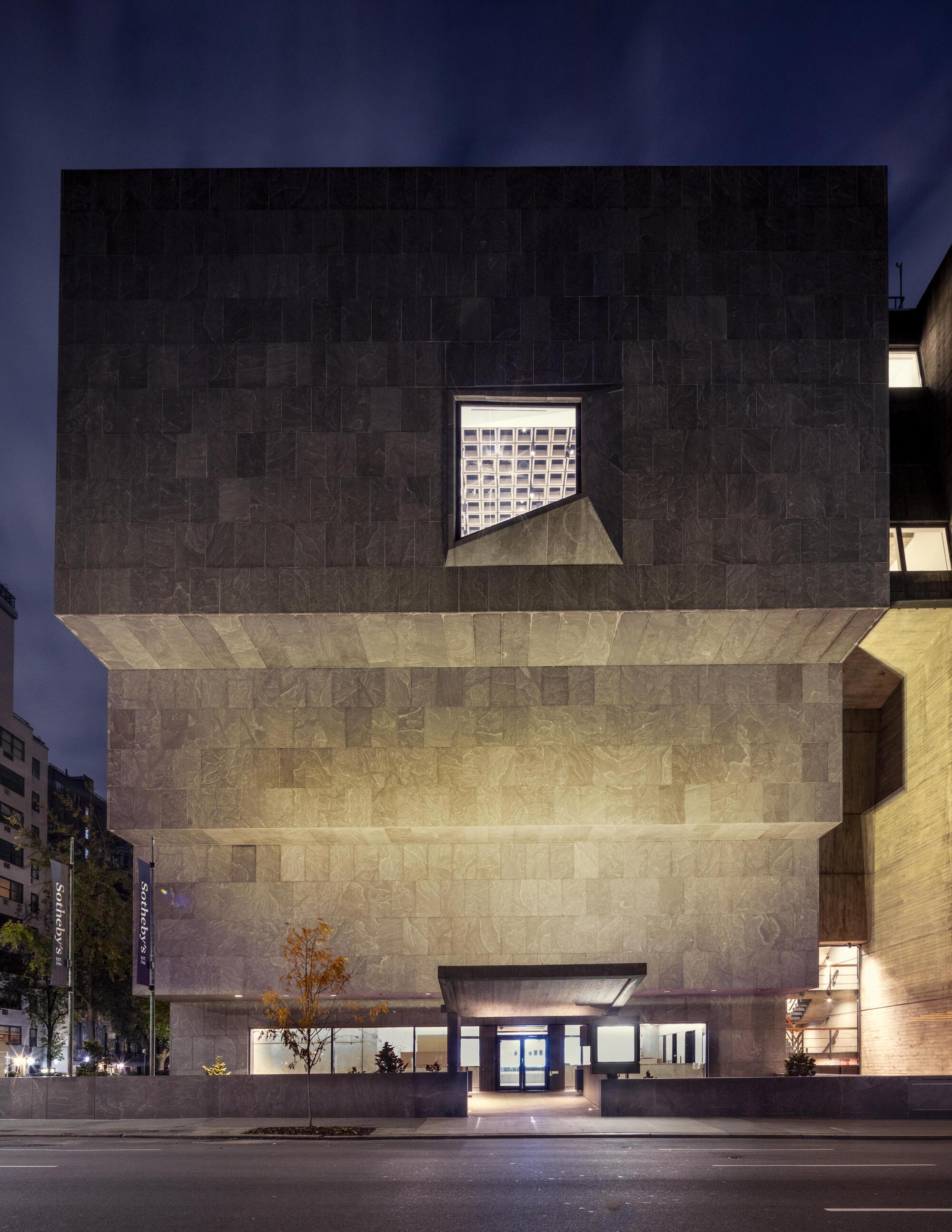 Frontal view of the inverted stepped stone facade with a single angled window, a small autumn tree and canopy entrance at night