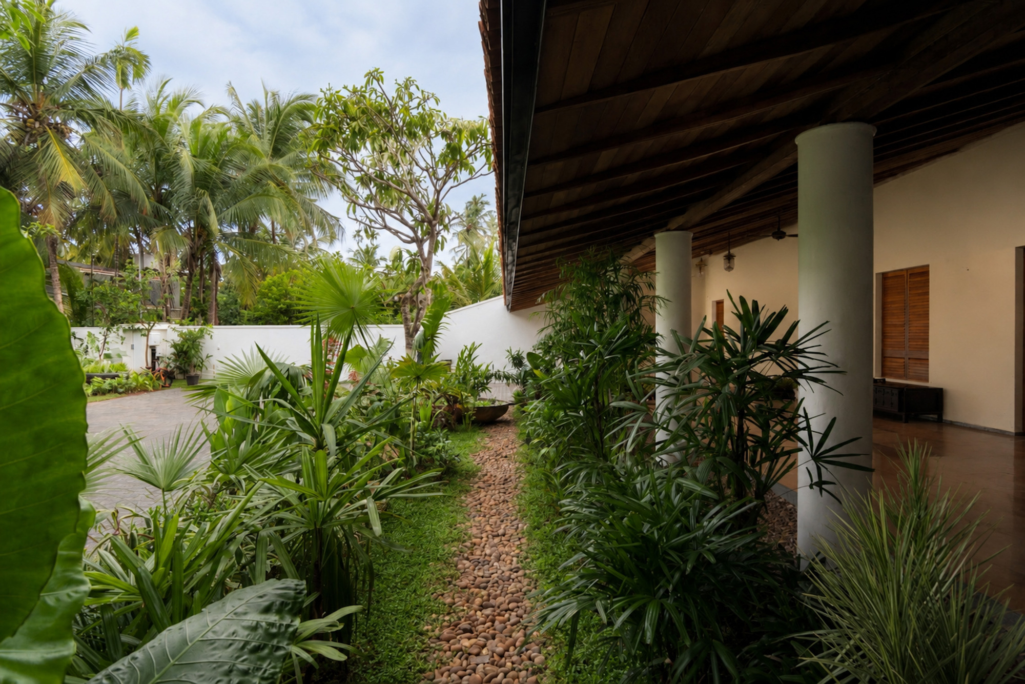 Pebble path running between tropical planting beside a veranda with white columns and a dark timber ceiling under overcast light