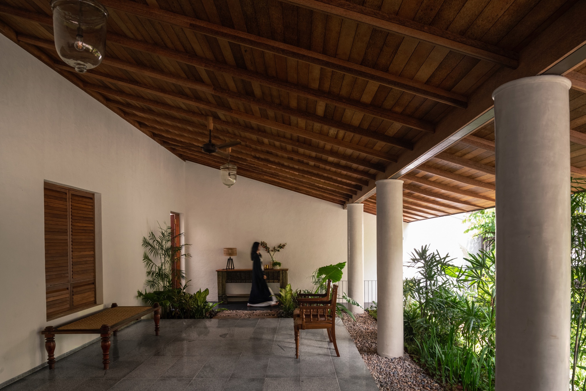 Covered veranda with round white columns, timber rafters and a woman walking past a console table, with gravel and plants beside it