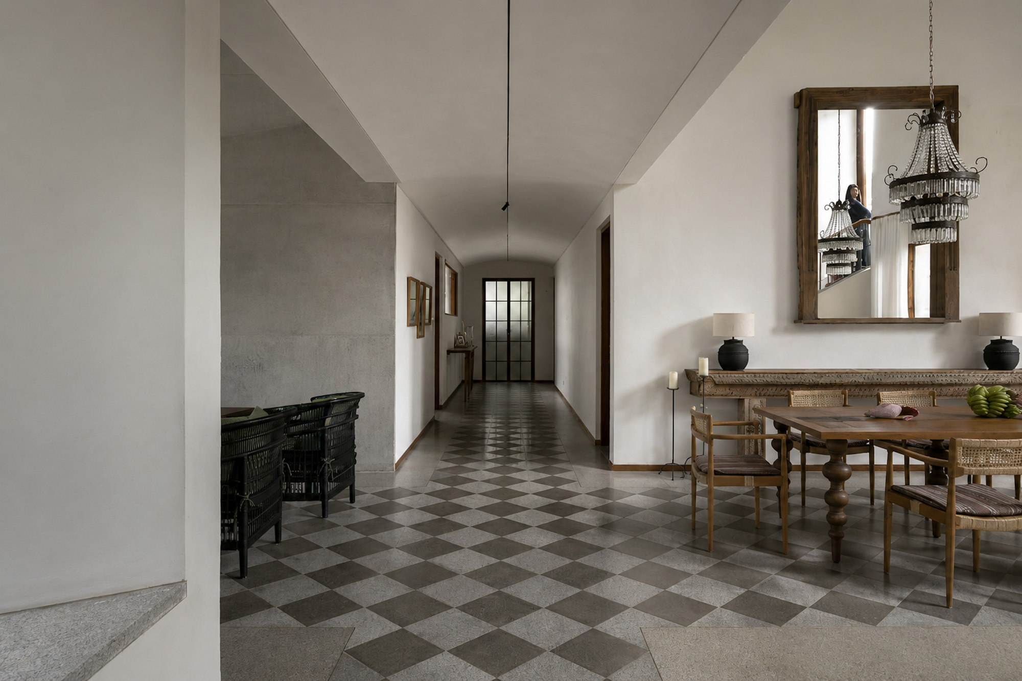 Hallway with checkerboard stone floor, vaulted ceiling and a dining table beneath a crystal chandelier, a woman reflected in the mirror