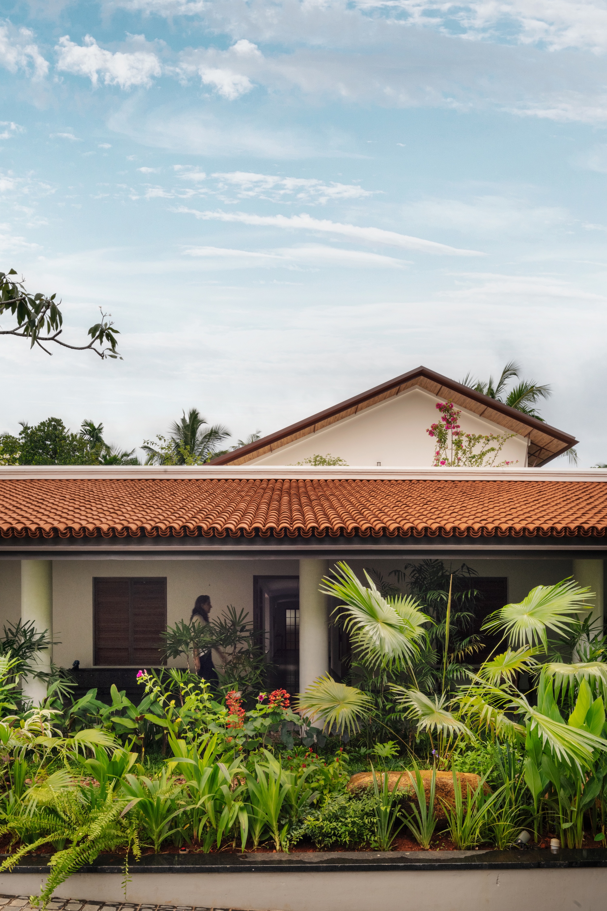 Vertical view of the tiled roof and gabled upper volume above a planted bed of palms and red flowers under a pale sky