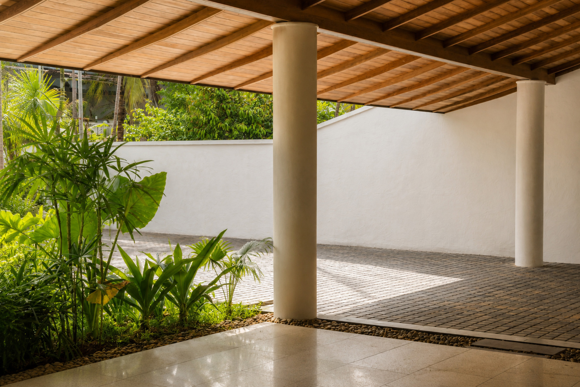 Covered carport with round white columns and timber ceiling, opening to a tropical planting bed and brick paved court in dappled light