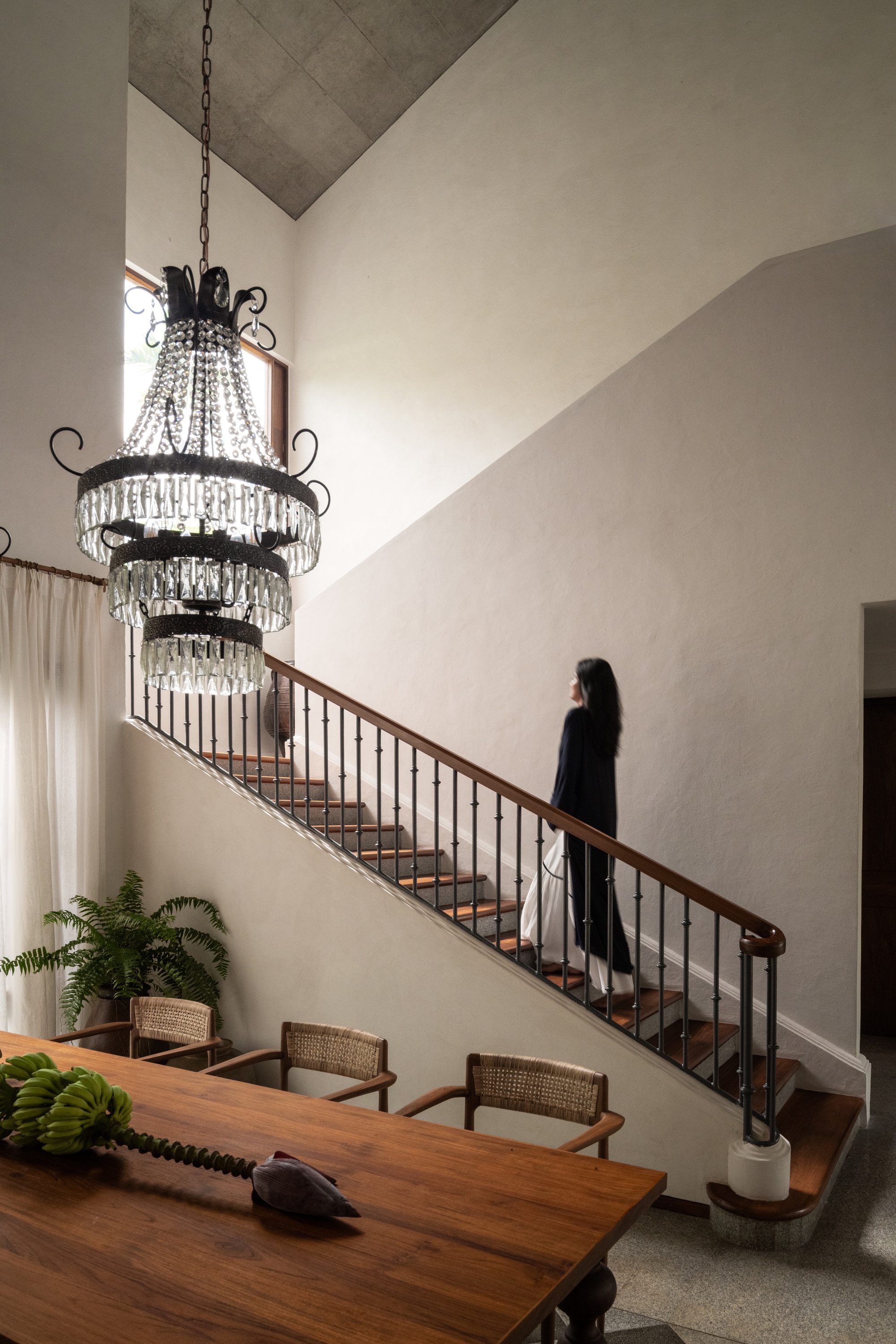 Woman climbing a timber and iron staircase beside a crystal chandelier and dining table, with a high concrete ceiling above
