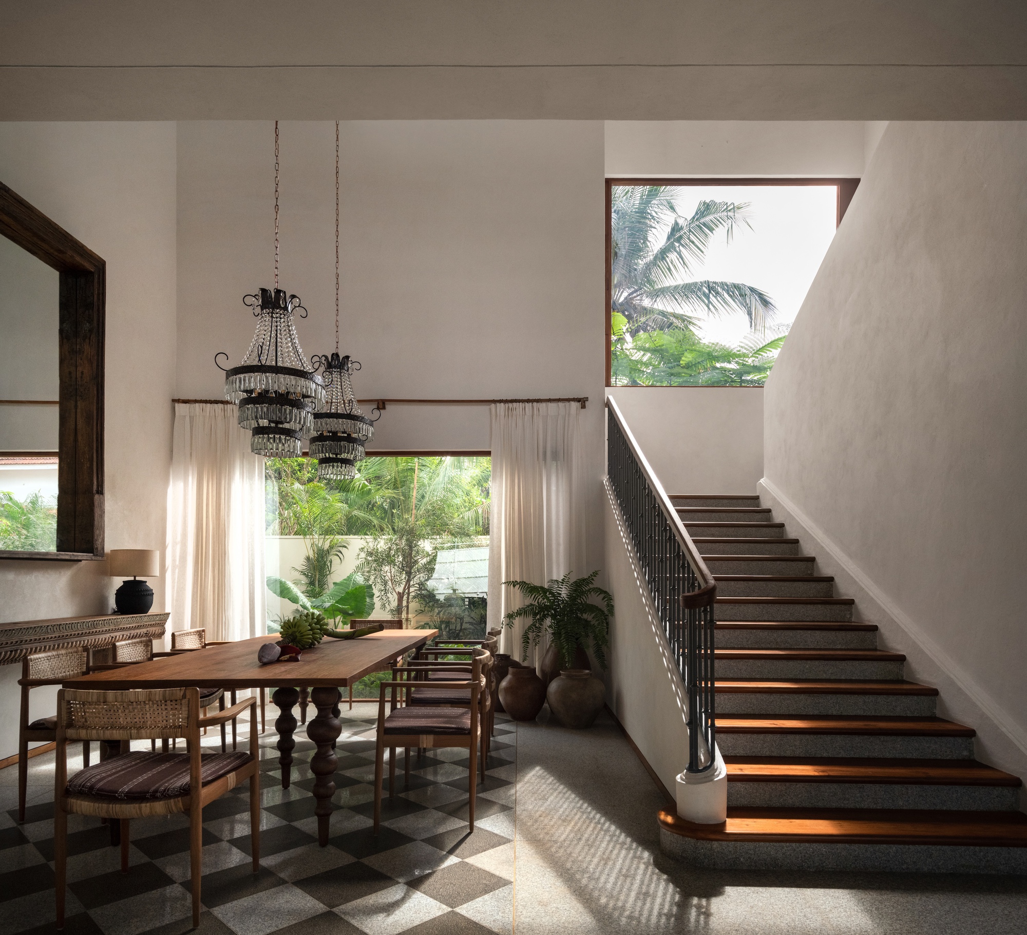 Dining area with checkerboard floor and twin chandeliers beside a terrazzo and timber stair, with palm fronds in the window beyond