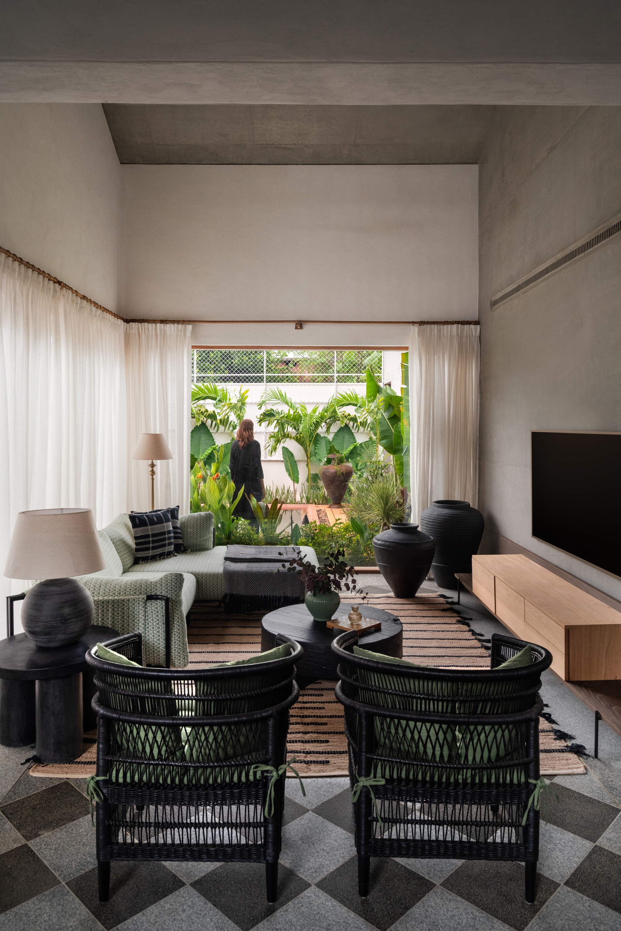 Living room with two black woven chairs on a checkered stone floor, facing sheer curtains and a garden with a person outside